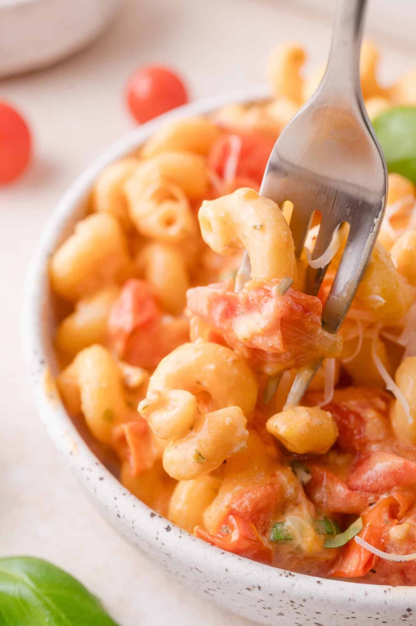 A fork lifting a portion of creamy Boursin pasta with diced tomatoes from a speckled bowl, with cherry tomatoes and basil leaves nearby.
