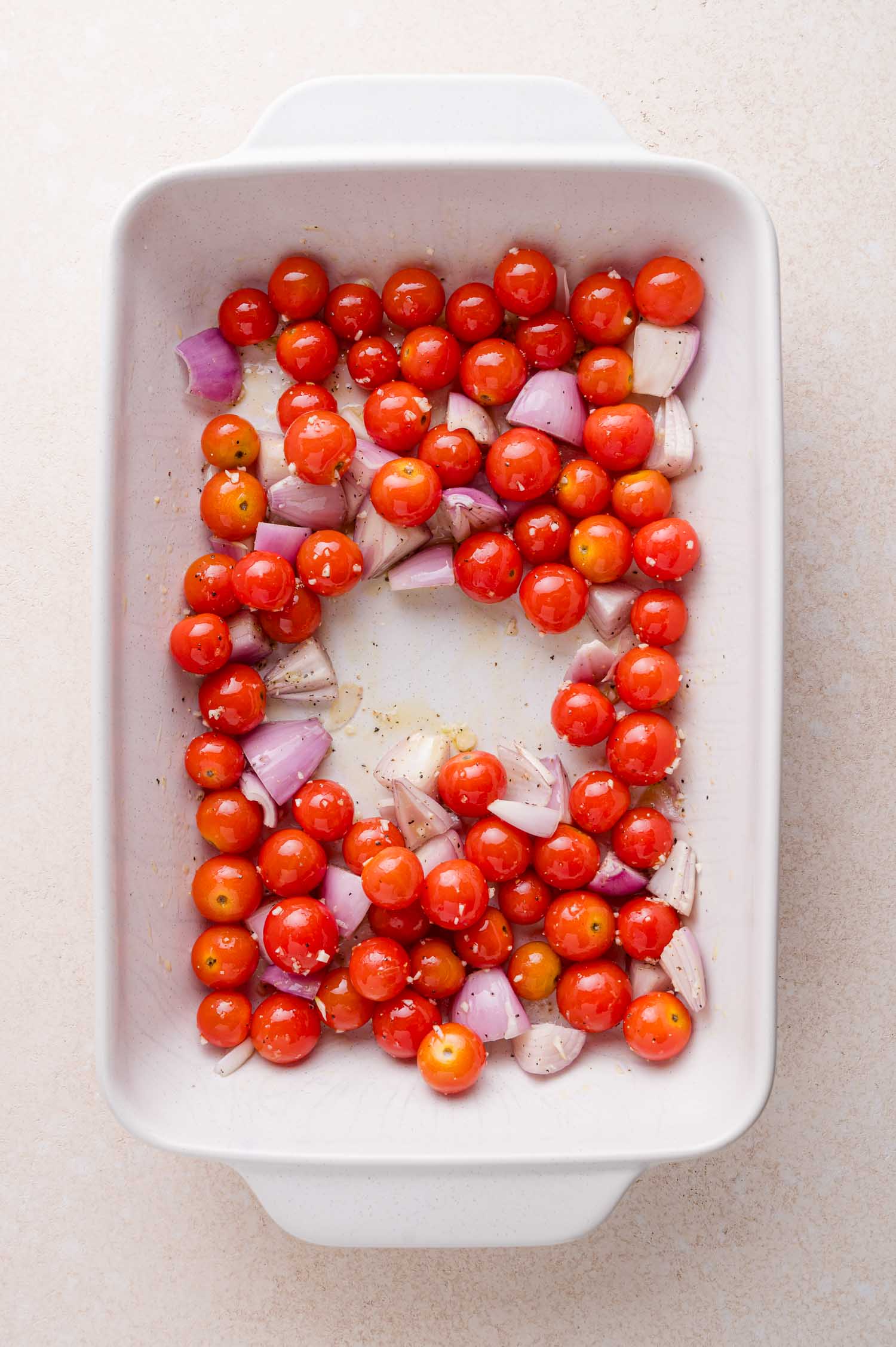 Cherry tomatoes and chopped red onions seasoned with salt, pepper, and oil in a white baking dish.