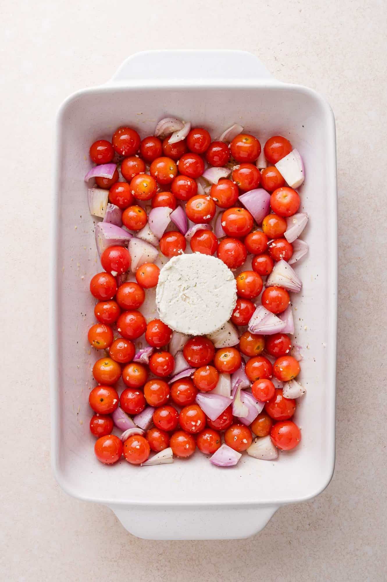 A white baking dish filled with cherry tomatoes, chopped red onions, garlic cloves, and a round block of white cheese in the center.