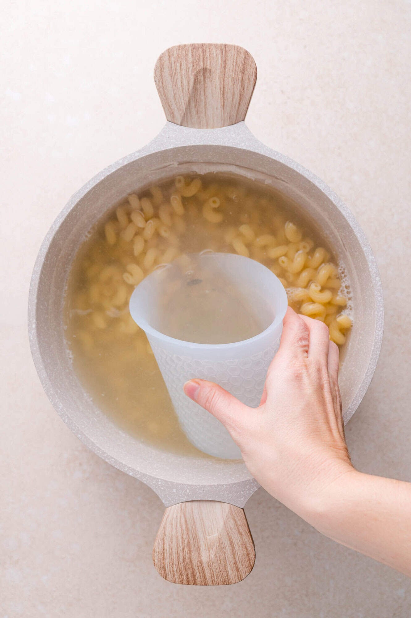 A hand holds a silicone measuring cup, scooping pasta water from a pot of cooked pasta on a light countertop.