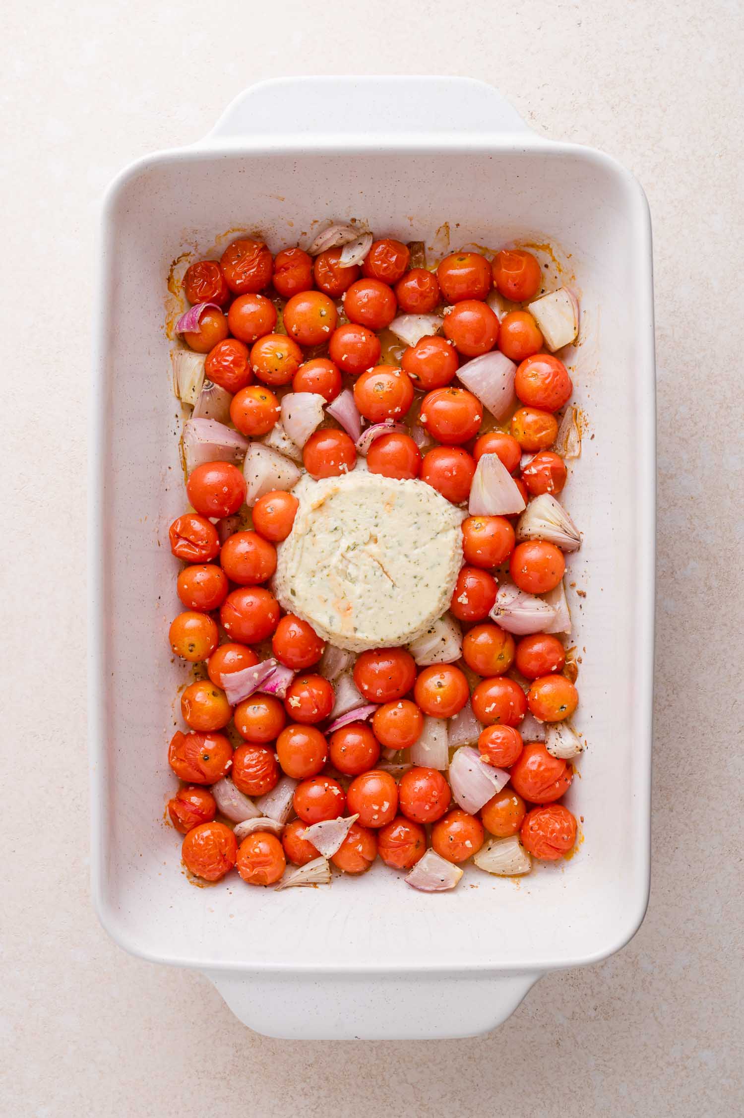 A baking dish filled with cherry tomatoes and chopped shallots, with a round block of cheese placed in the center.