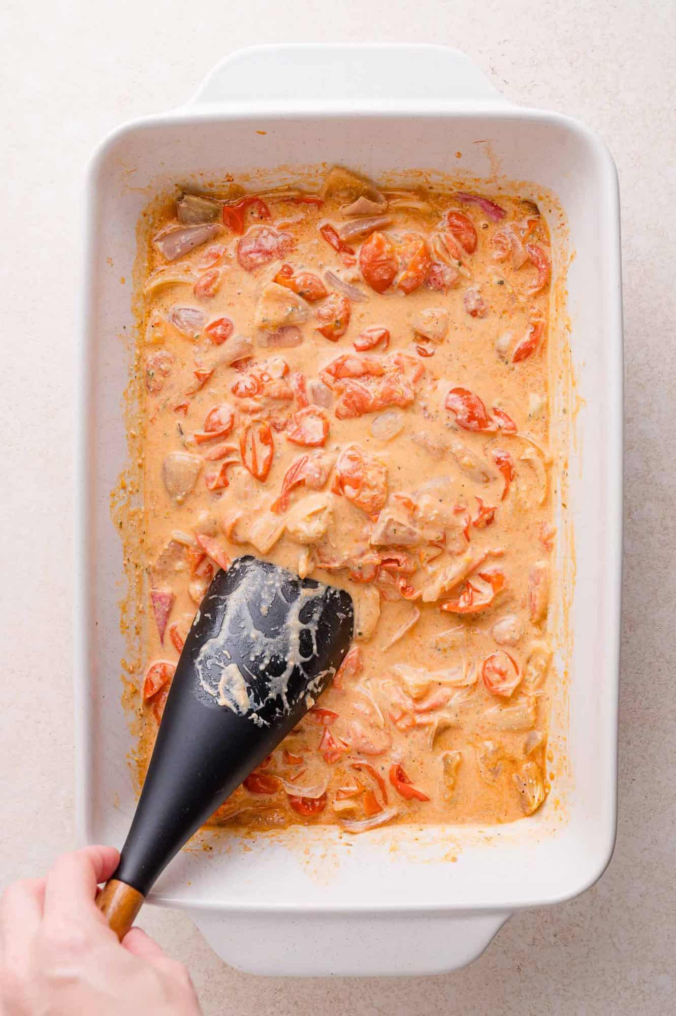A hand holds a black spatula stirring a creamy tomato and cheese mixture in a white rectangular baking dish.