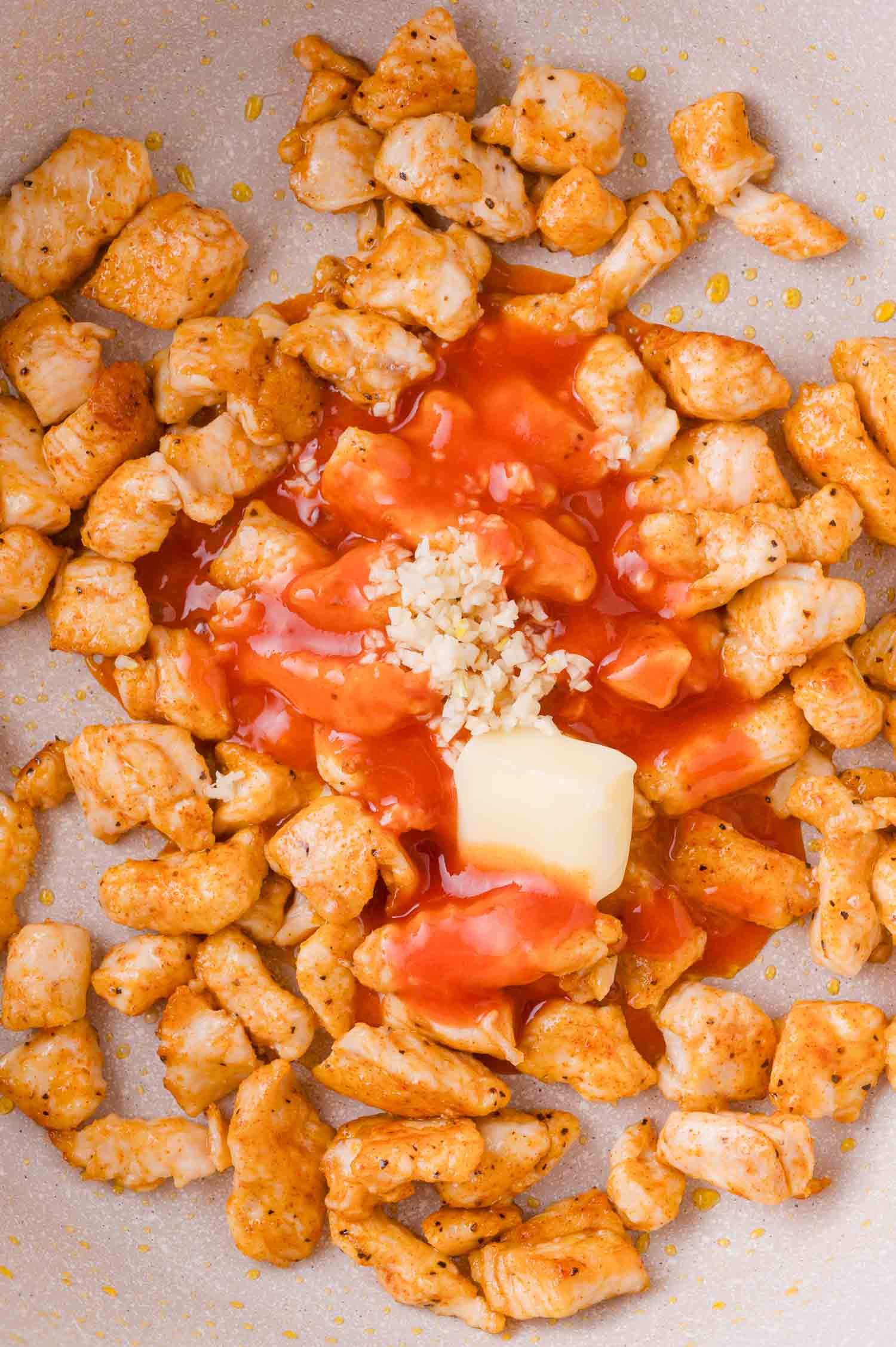 Seasoned, cooked chicken pieces in a pan with hot sauce, minced garlic, and a pat of butter.