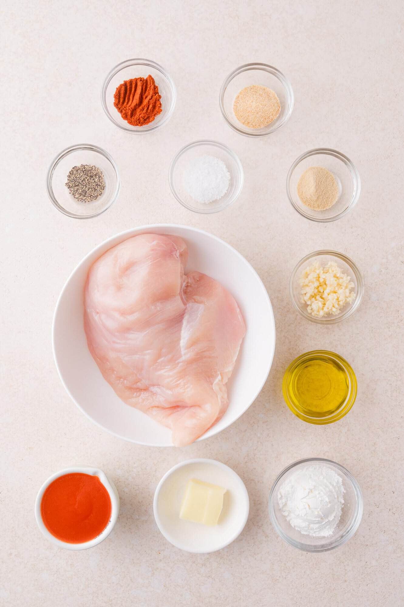 Raw chicken breast in a white bowl surrounded by small bowls of spices, garlic, oil, hot sauce, butter, and cornstarch on a light surface.