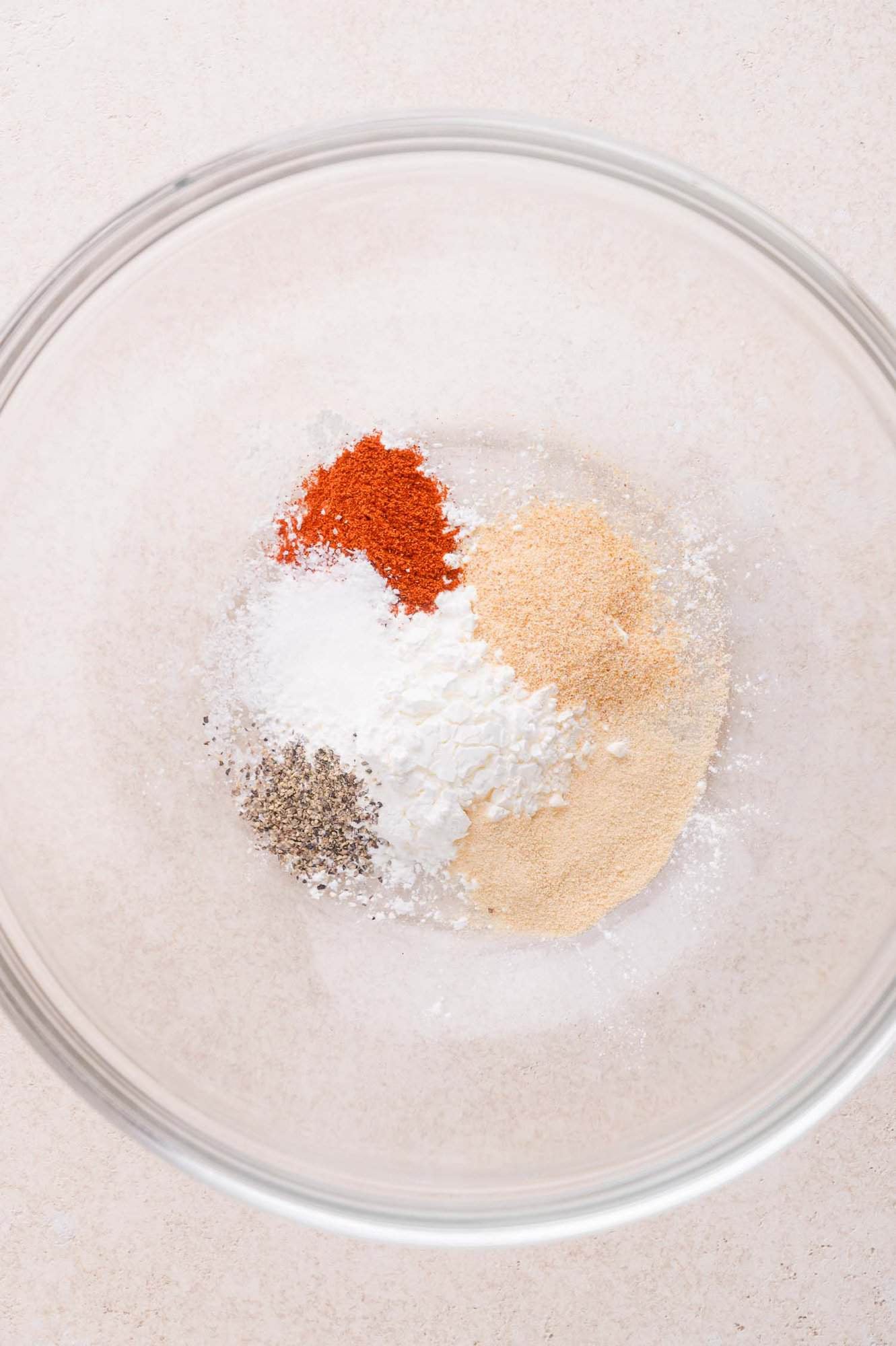 A clear glass bowl containing separate piles of paprika, black pepper, salt, cornstarch, garlic powder, and onion powder on a light surface.