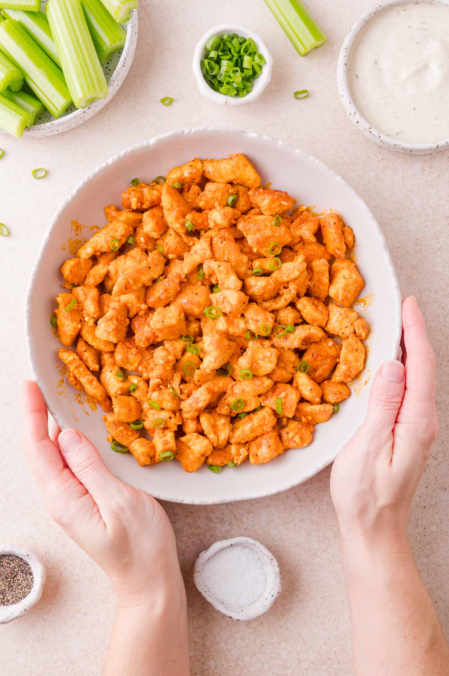 A bowl of buffalo chicken bites garnished with green onions, surrounded by celery sticks, ranch dressing, and seasonings, with two hands holding the bowl.