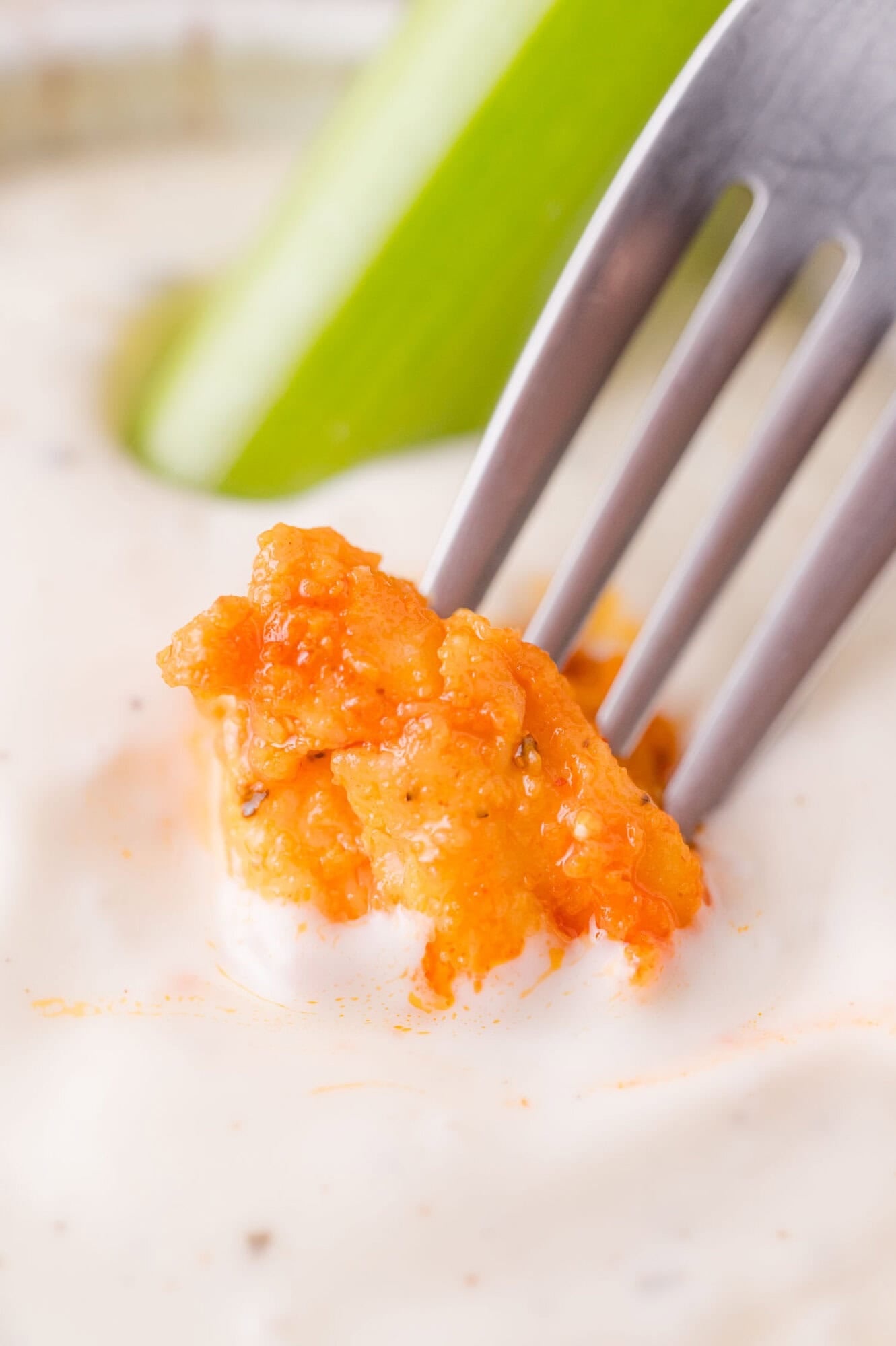 A fork holds a piece of buffalo chicken dipped in creamy ranch dressing, with a celery stick in the background.