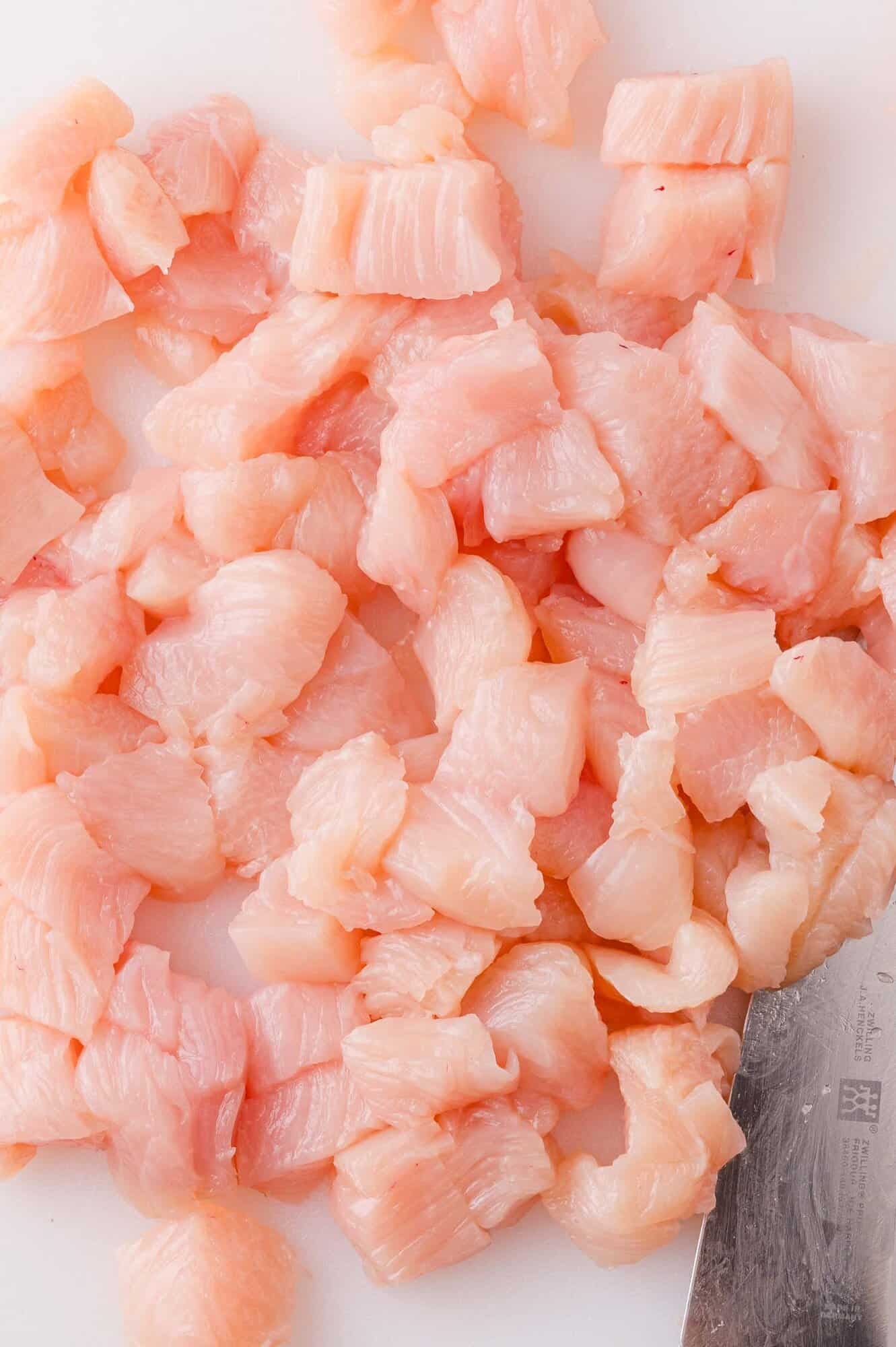Diced raw chicken breast pieces on a white cutting board with a metal knife visible in the corner.