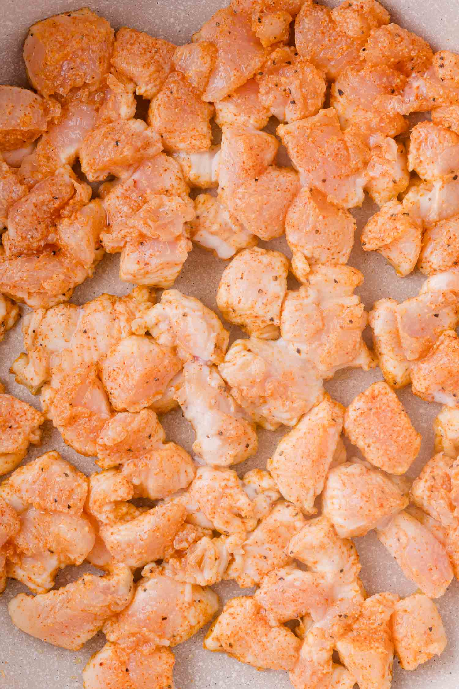 Raw, diced chicken breast coated with spices spread out in a single layer on a light-colored surface, ready to be cooked.