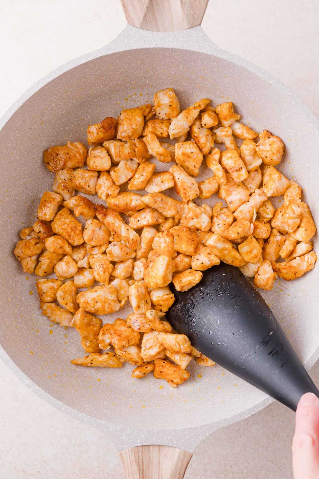 Diced, seasoned chicken cooking in a light-colored skillet is being stirred with a black spatula.