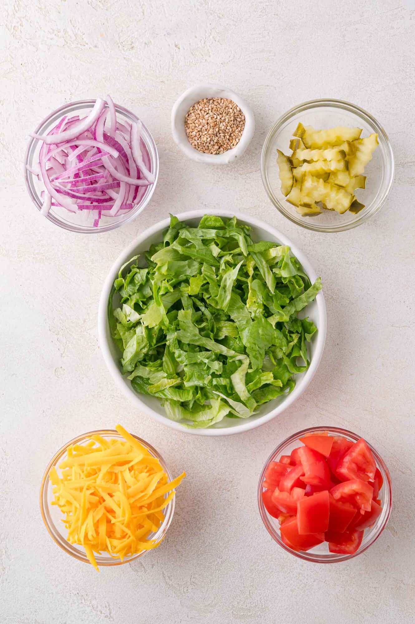 Five bowls containing shredded lettuce, diced tomatoes, shredded cheddar cheese, sliced pickles, and sliced red onions, with a small bowl of sesame seeds in the center background.