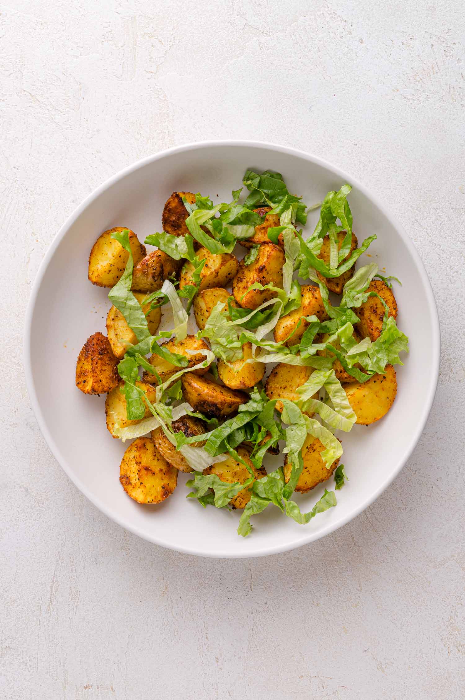 A white bowl containing roasted potato slices garnished with chopped lettuce on a light-colored surface.