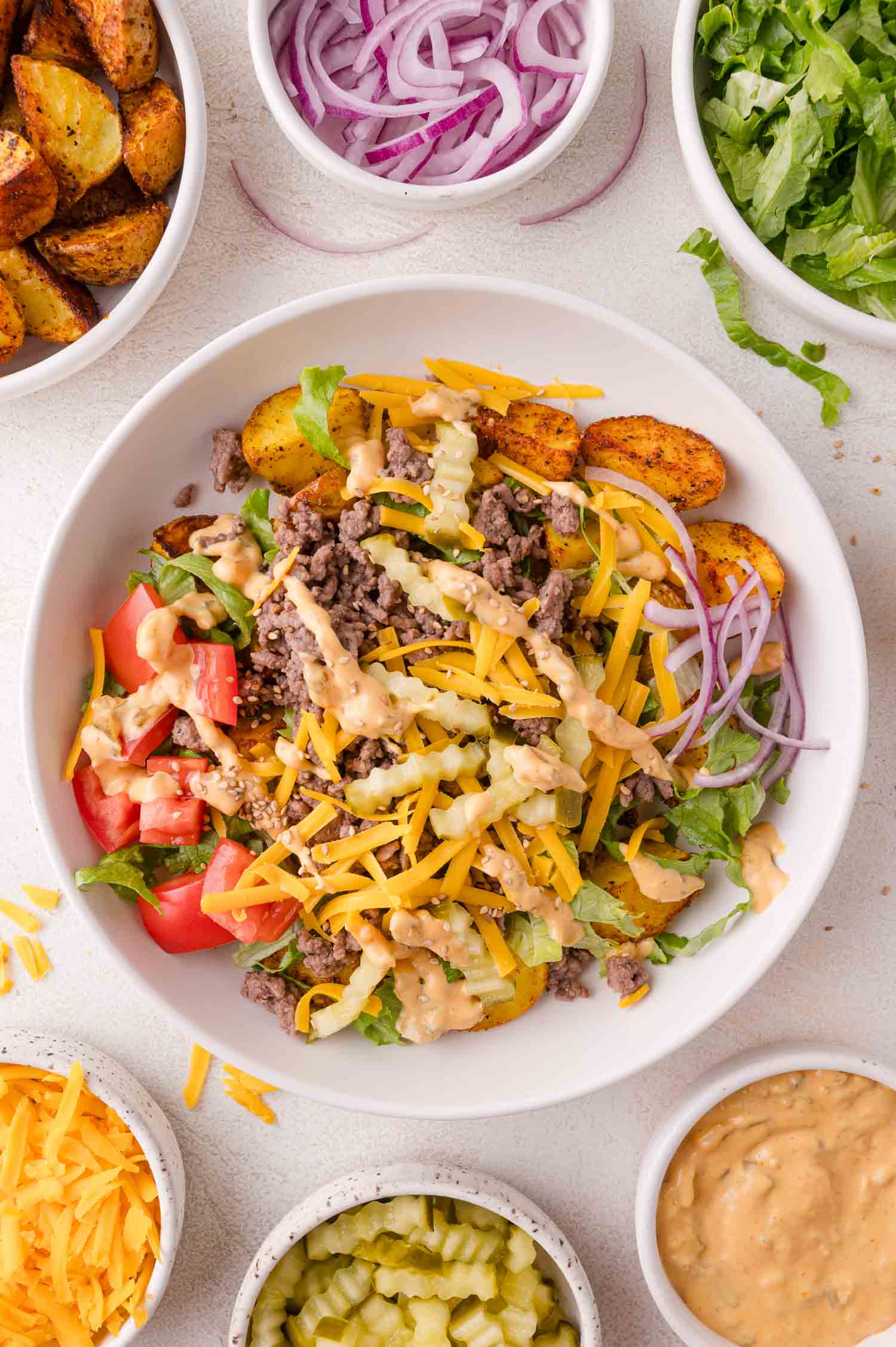 A burger bowl with ground beef, shredded cheese, lettuce, tomato, red onion, and sauce, surrounded by small dishes of potatoes, lettuce, red onion, pickles, shredded cheese, and sauce.