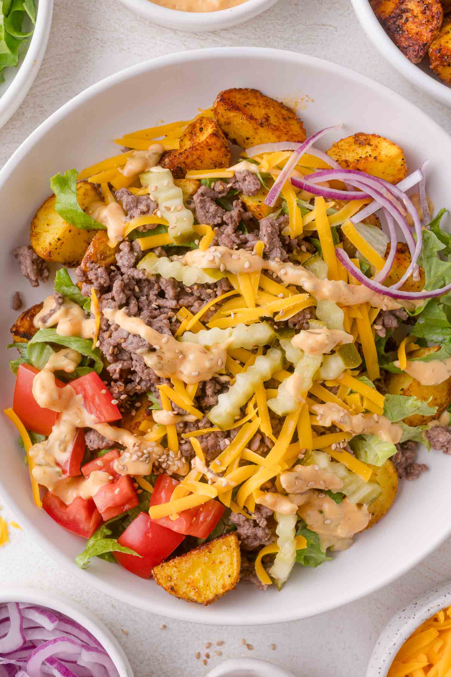 A bowl filled with ground beef, shredded cheddar cheese, lettuce, tomato, red onion, diced pickles, seasoned potato wedges, and a drizzle of creamy dressing.