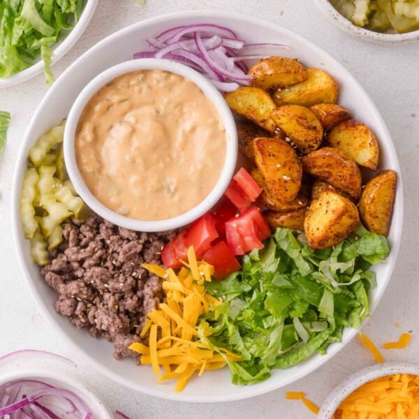 A burger bowl filled with ground beef, diced tomatoes, shredded cheddar, lettuce, sliced red onions, pickles, roasted potato wedges, and a serving of creamy sauce.