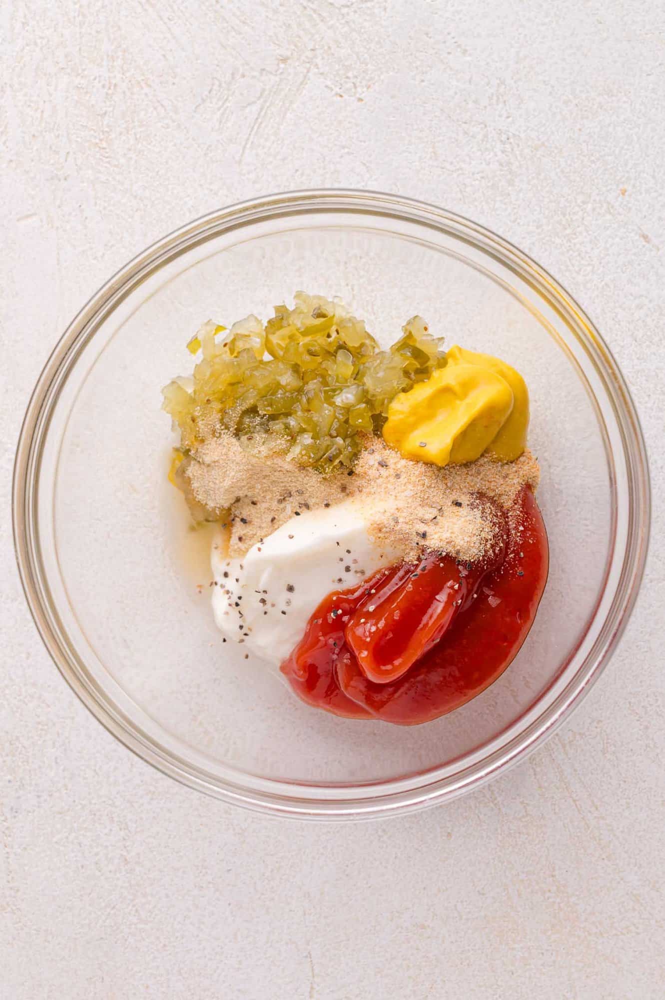 A clear glass bowl containing ketchup, mustard, mayonnaise, sweet relish, black pepper, and seasonings on a white surface.
