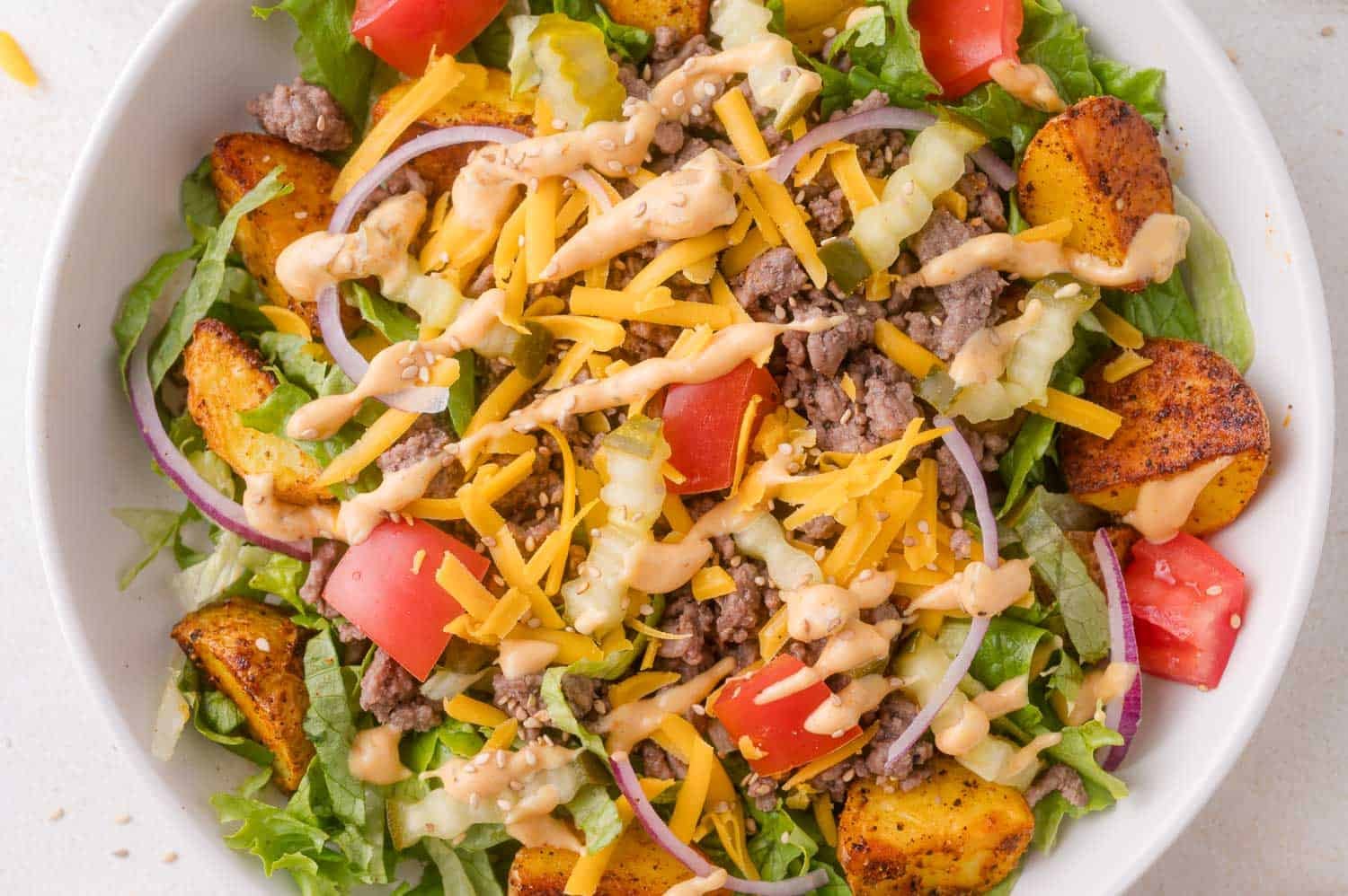 Burger bowl with ground beef, cheddar cheese, tomato, red onion, lettuce, pickles, roasted potatoes, and a drizzle of creamy dressing.