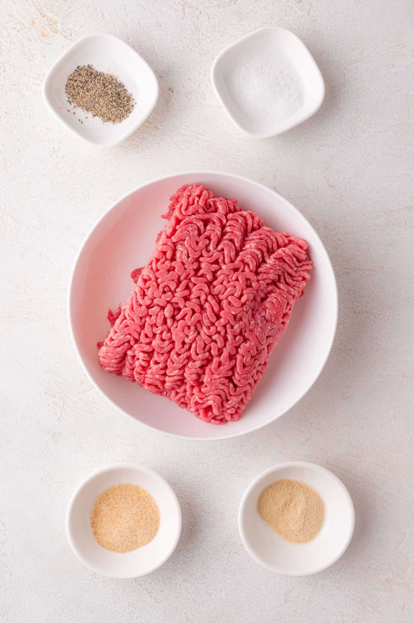 Raw ground beef in a white bowl surrounded by small dishes containing black pepper, salt, garlic powder, and onion powder on a light surface.