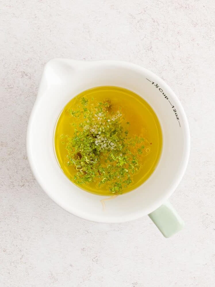 A white measuring cup filled with olive oil, lime zest, salt, and pepper on a light surface.