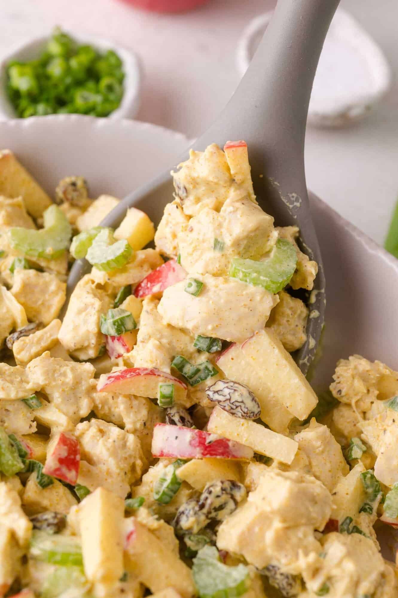 A close-up of a spoon scooping curried chicken salad with apple slices, celery, raisins, and green onions from a bowl.