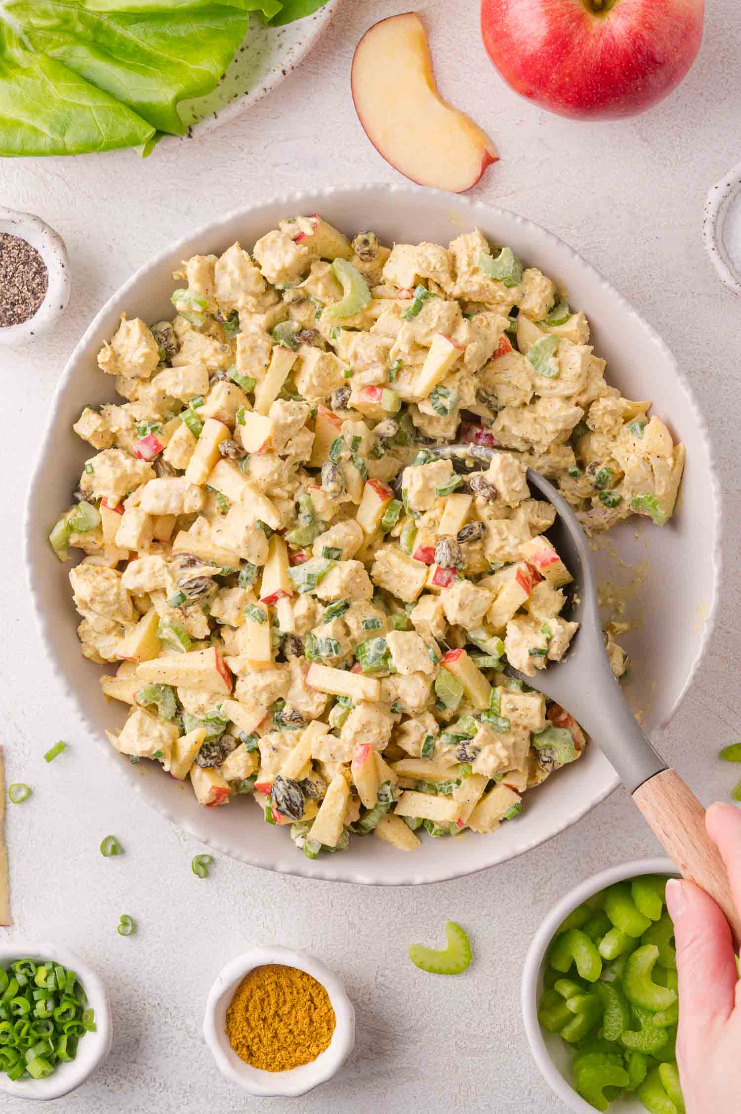 A bowl of chicken salad with diced apples, celery, raisins, and green onions, with a spoon in the bowl. Surrounding ingredients include apple slices, celery, lettuce, and spices.