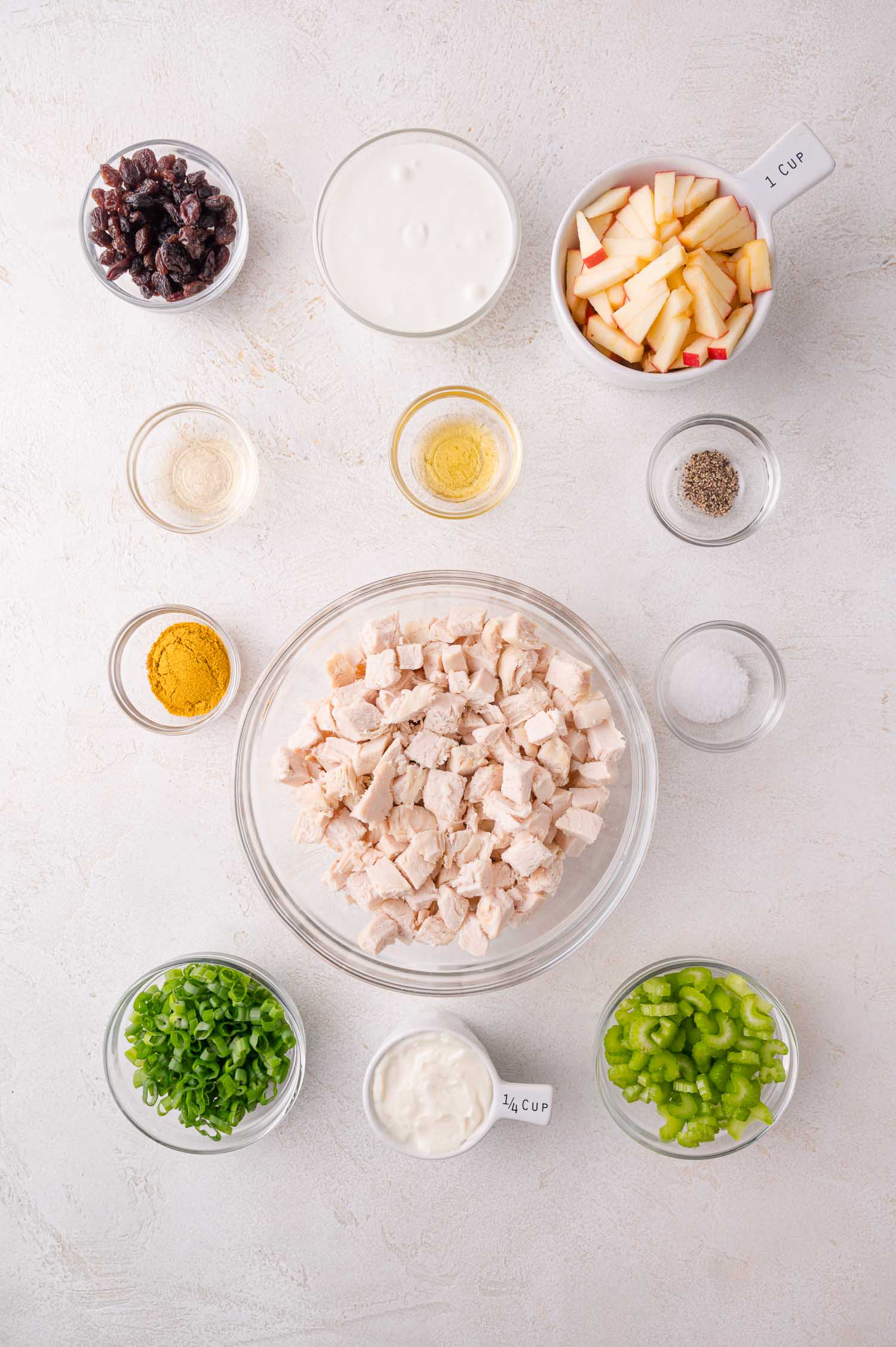 Overhead view of ingredients for a recipe, including diced chicken, chopped celery, green onions, apple pieces, yogurt, mayonnaise, curry powder, raisins, vinegar, and seasonings in bowls.