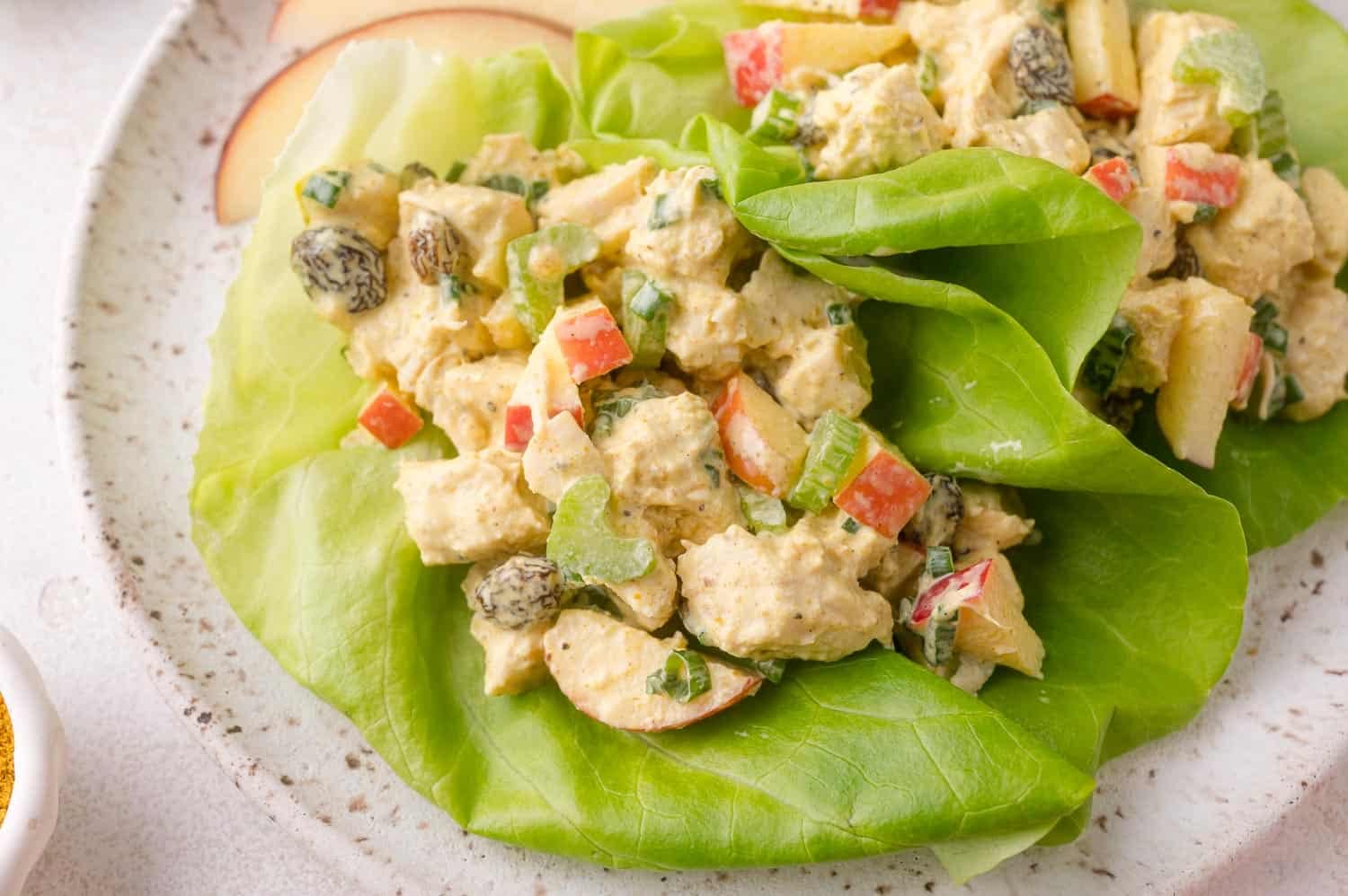 Chicken salad with diced celery, raisins, and greens served on large lettuce leaves on a speckled plate.