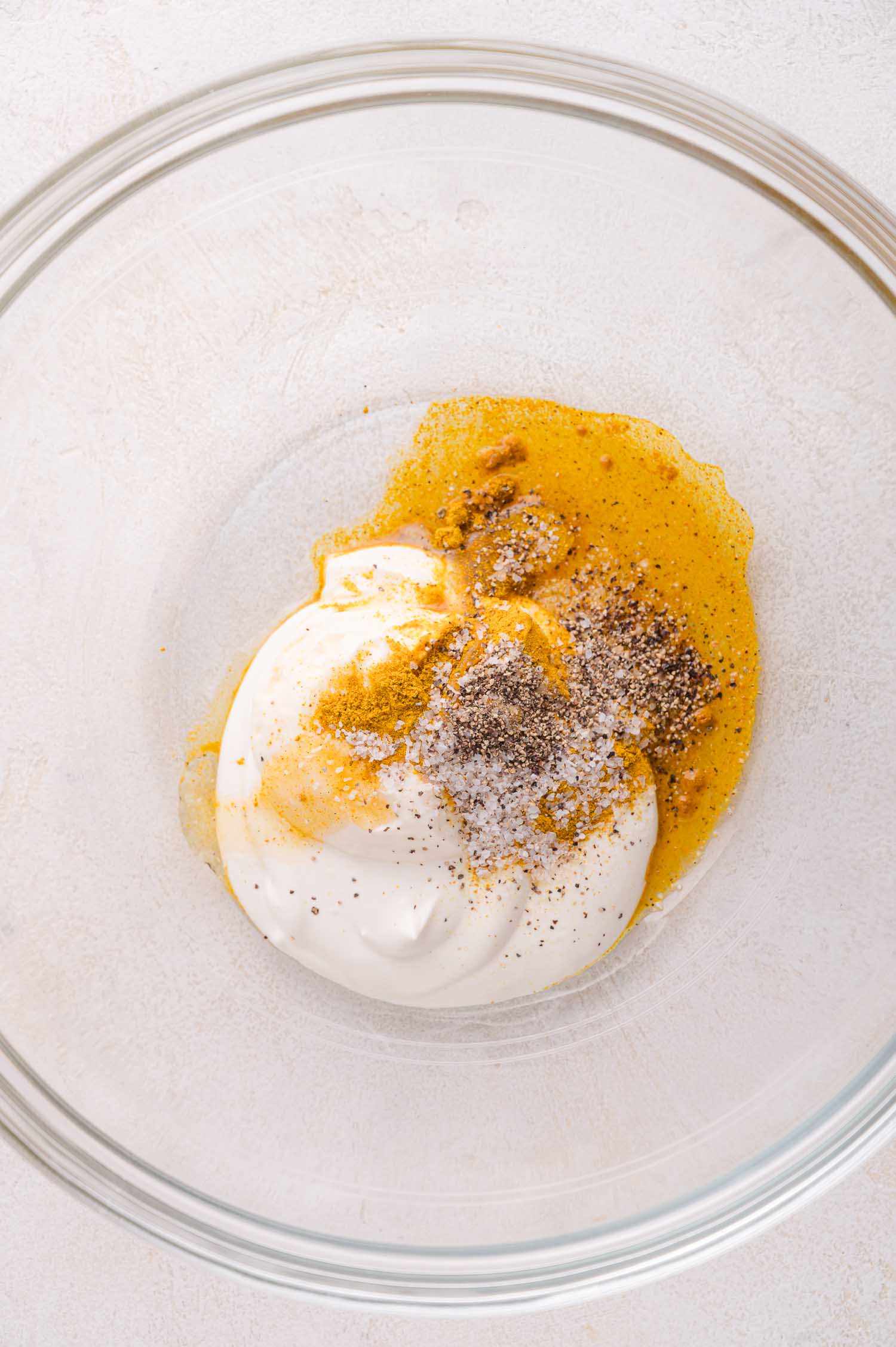 A glass bowl contains mayo, mustard, black pepper, salt, and spices, ready to be mixed together.