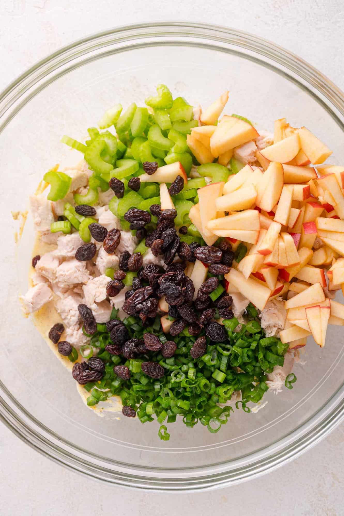 A glass bowl containing chopped celery, diced apples, raisins, green onions, and chunks of cooked chicken.
