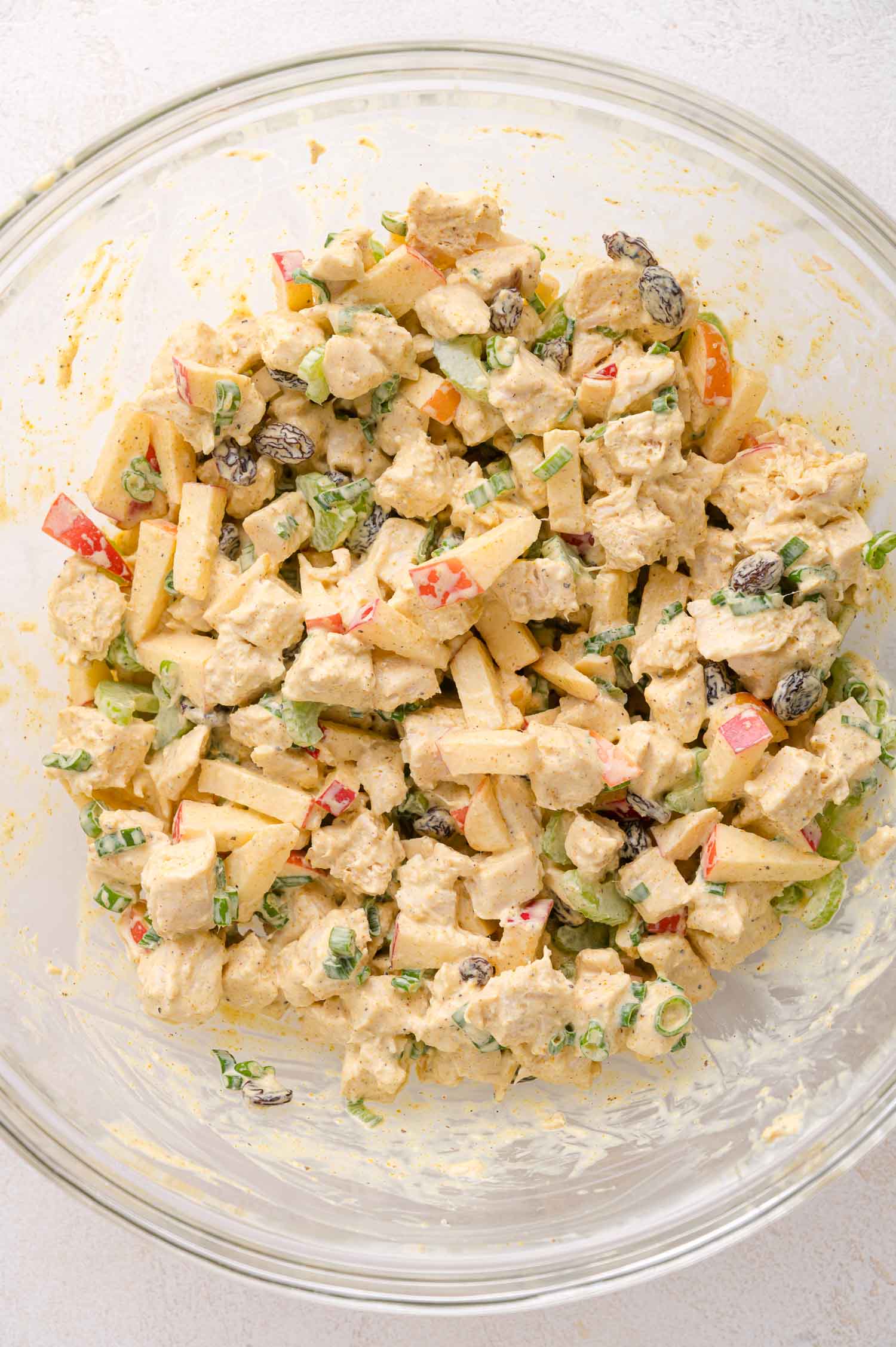 A glass bowl filled with a mixed chicken salad containing diced chicken, apple slices, green onions, raisins, and a creamy dressing.