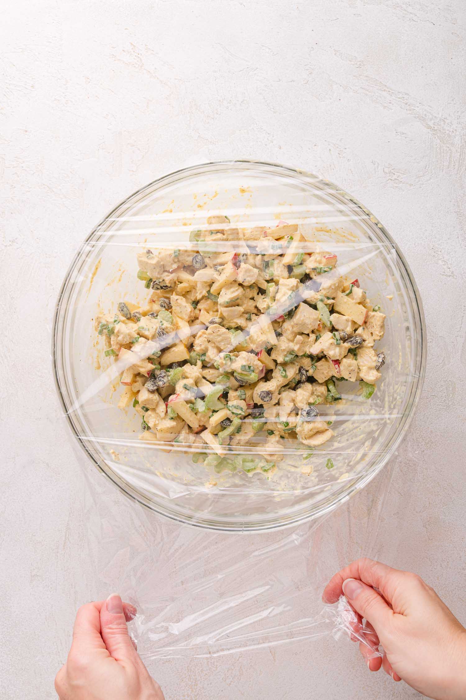 Hands cover a glass bowl of curry chicken salad with plastic wrap on a light-colored surface.