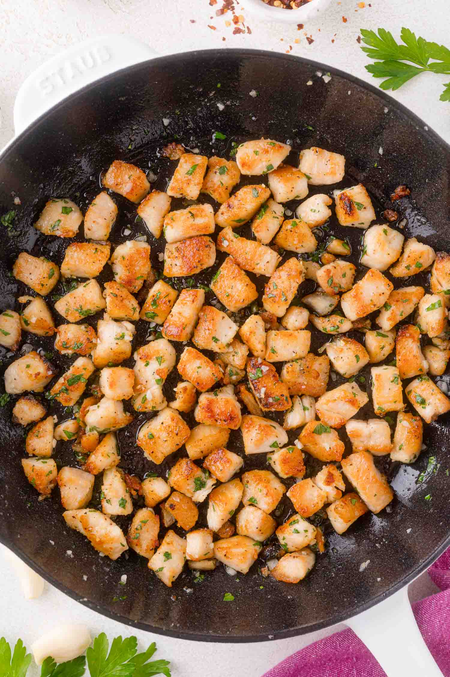 Diced, seasoned chicken pieces are pan-fried and browned in a black skillet, garnished with chopped herbs and garlic.