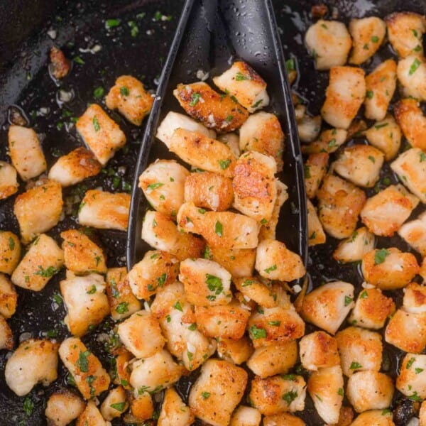 Spoon holding cooked, seasoned chicken bites in a skillet, garnished with chopped herbs and garlic.