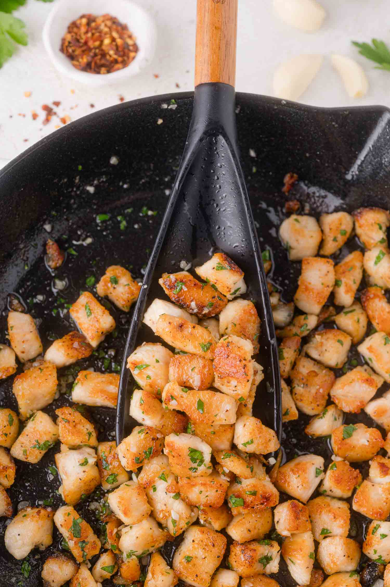Cubed pieces of seasoned chicken breast are cooked and browned in a skillet, with a spatula lifting some pieces. Chopped herbs are sprinkled on top.