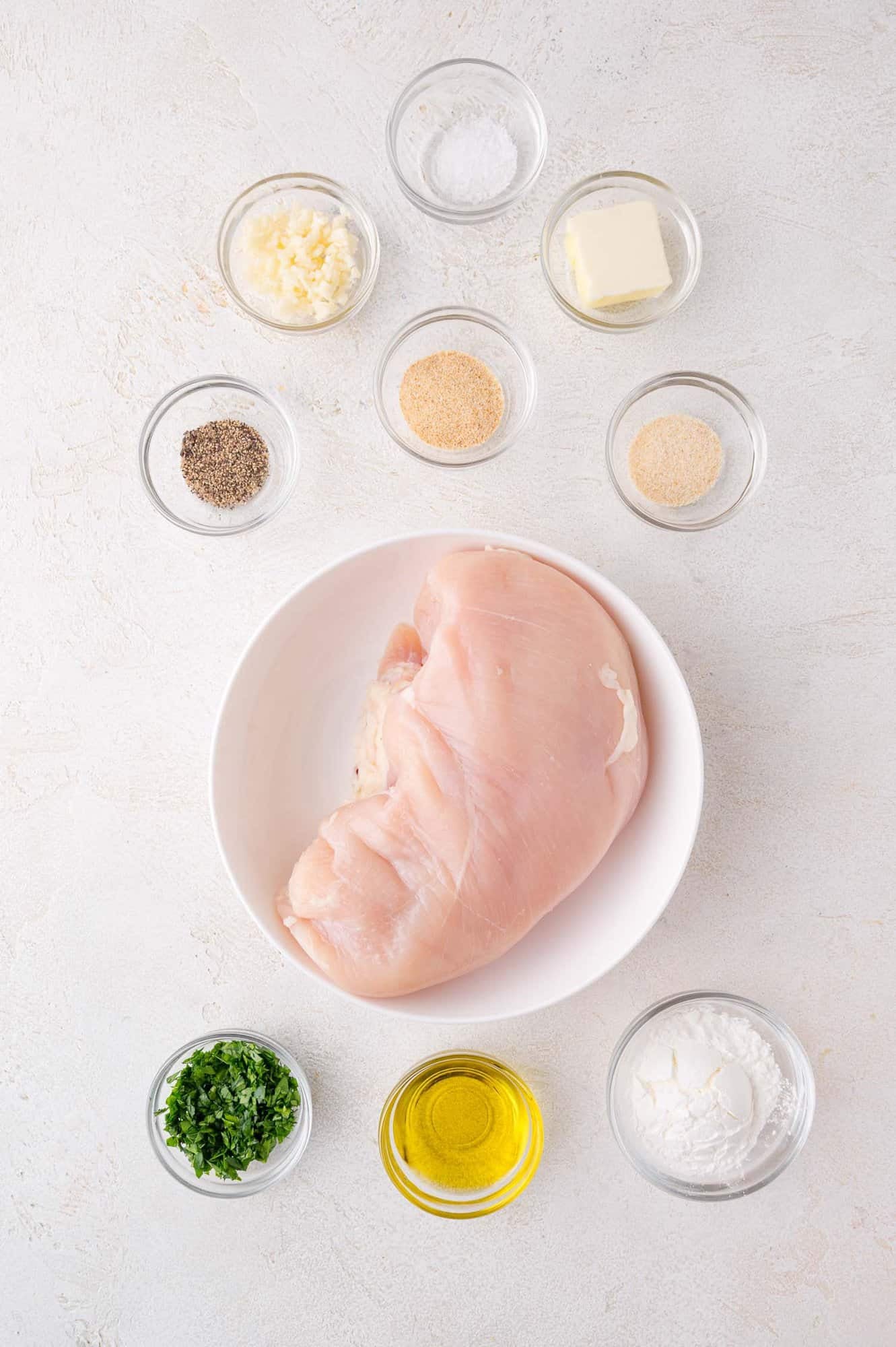 A raw chicken breast in a bowl surrounded by small bowls containing olive oil, butter, chopped herbs, minced garlic, black pepper, salt, and seasonings on a white surface.