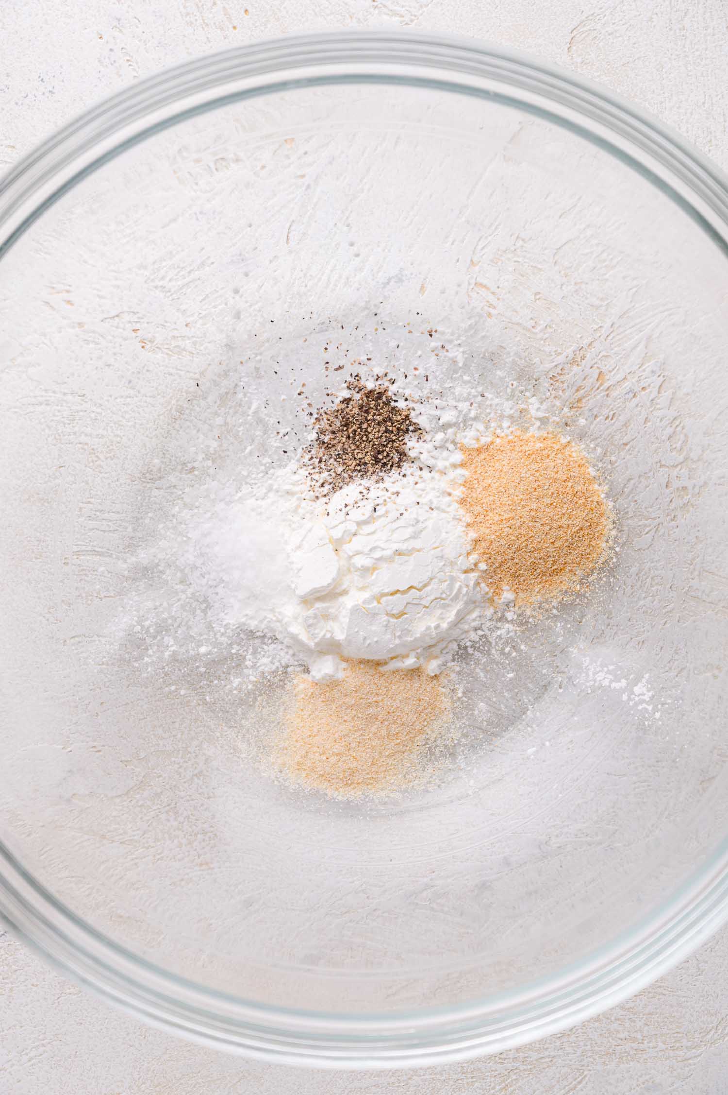 A glass bowl containing measured portions of cornstarch, garlic powder, and black pepper on a light surface.