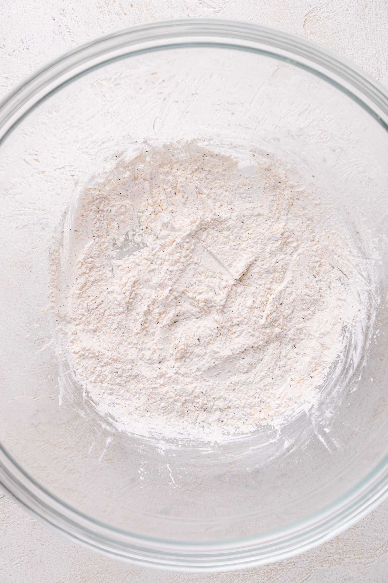 A clear glass bowl containing a mixture of dry, white cornstarch and seasonings on a light-colored surface.