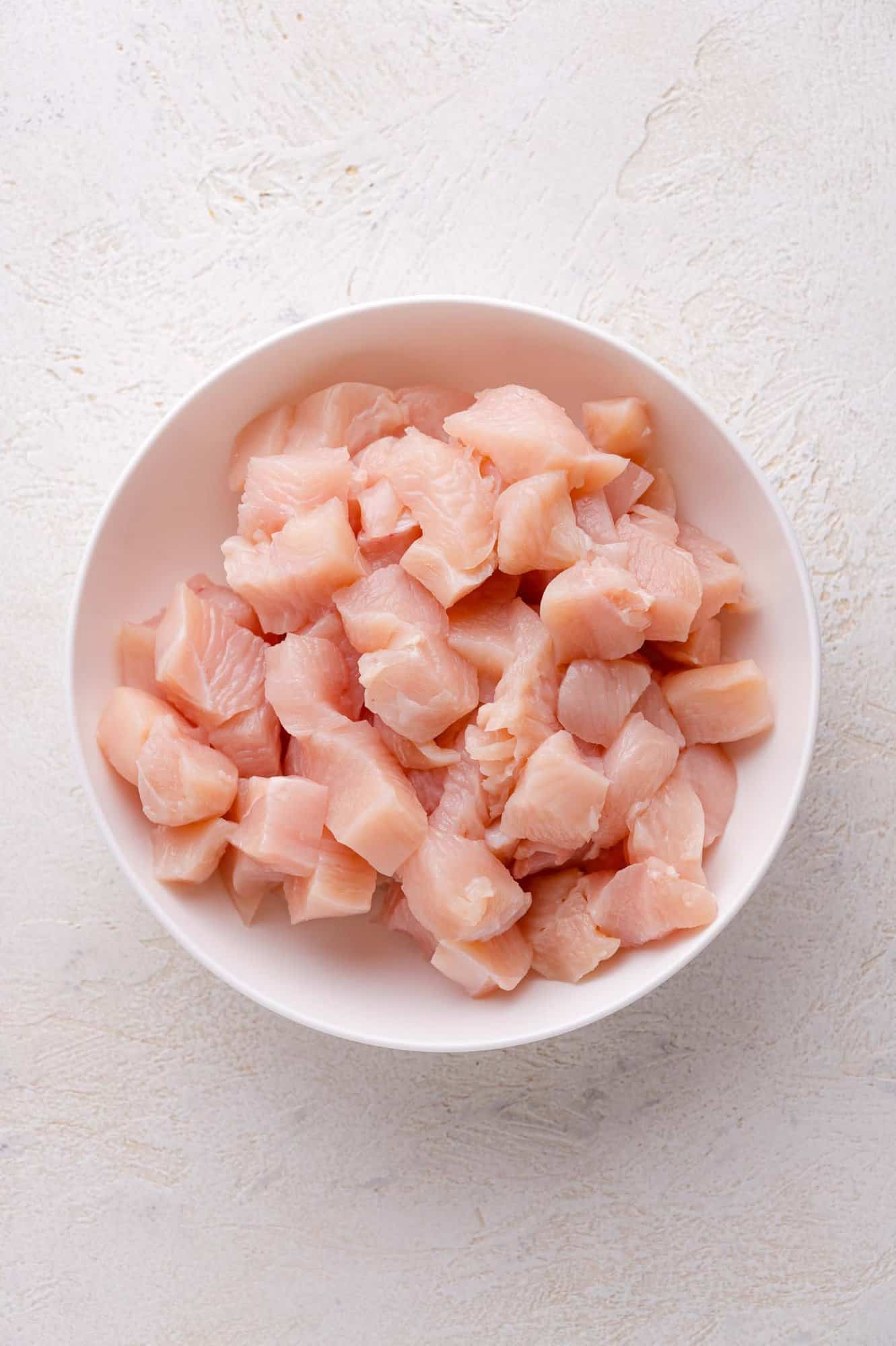 A white bowl filled with raw, diced chicken breast on a light textured surface.