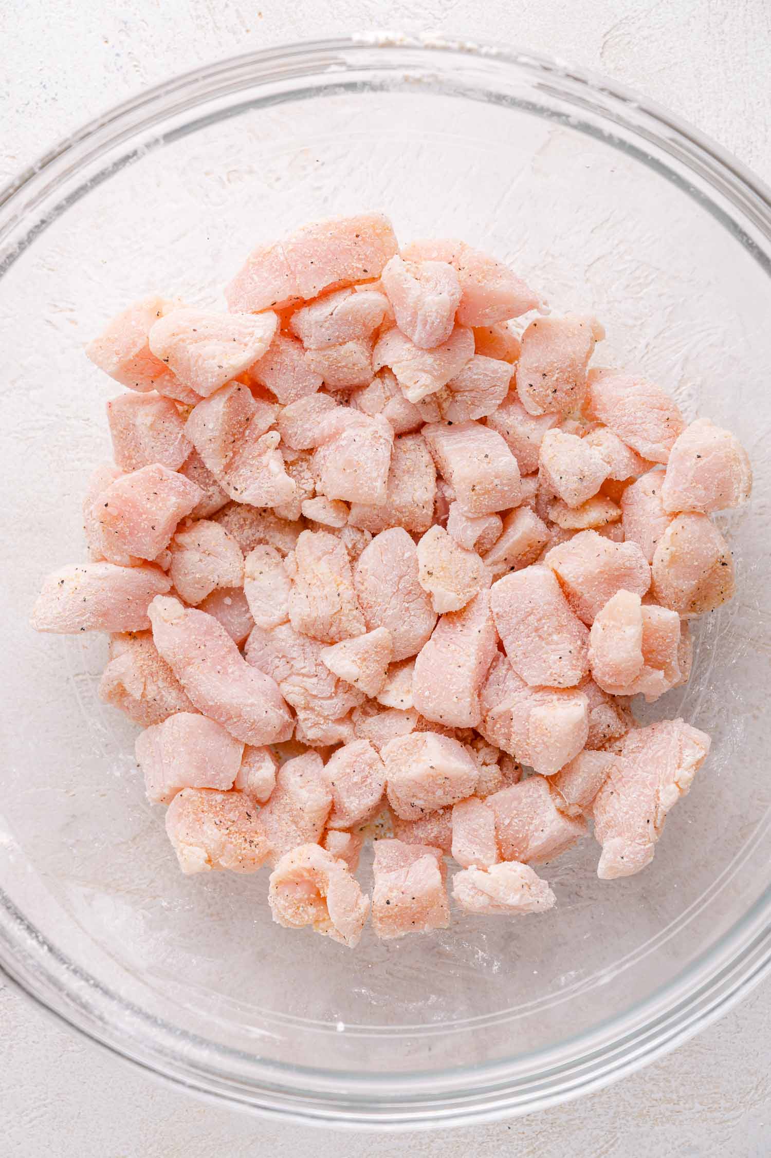 Cubed raw chicken pieces with seasoning in a clear glass bowl on a light-colored surface.