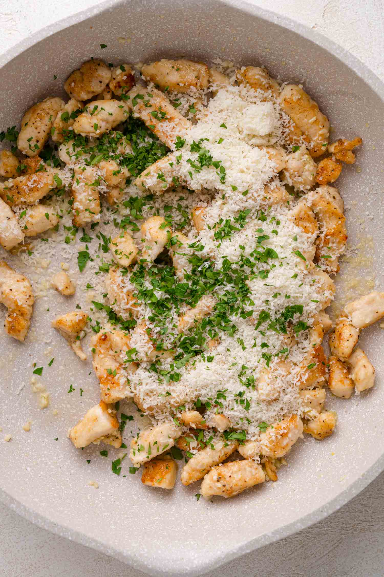 Cooked chicken pieces in a skillet topped with grated parmesan cheese and chopped parsley.
