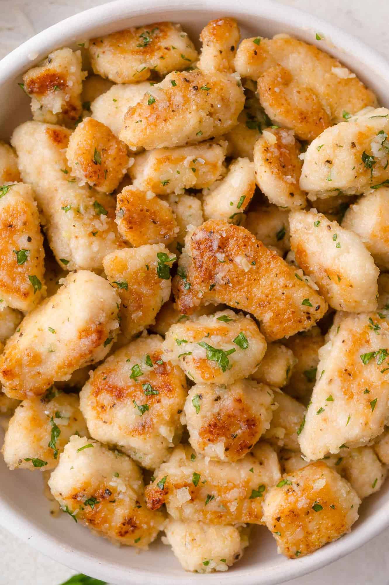 A bowl filled with golden-brown, pan-seared pieces of chicken garnished with parmesan and chopped herbs.