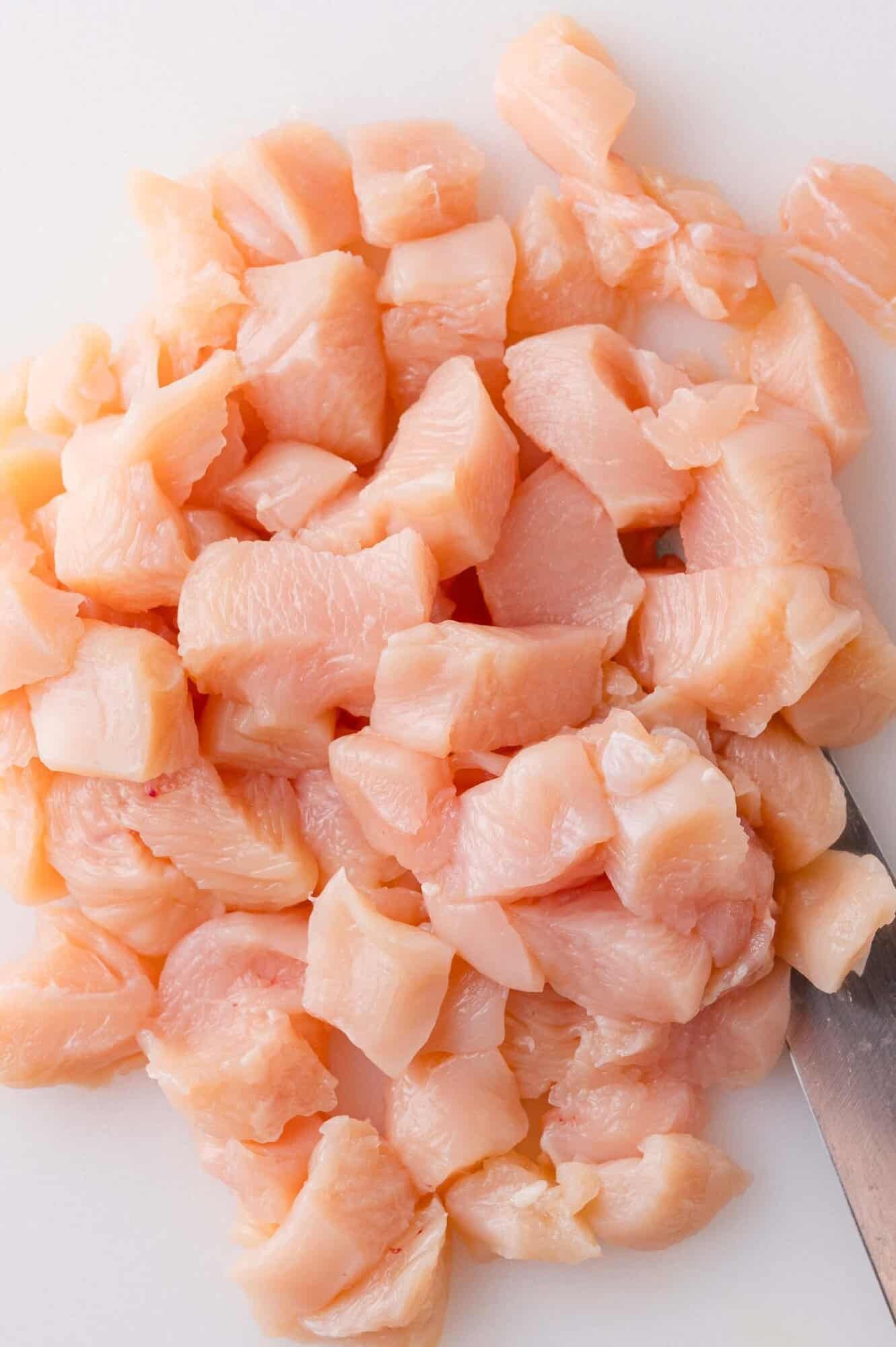 Diced raw chicken pieces on a white cutting board with a knife visible beside them.