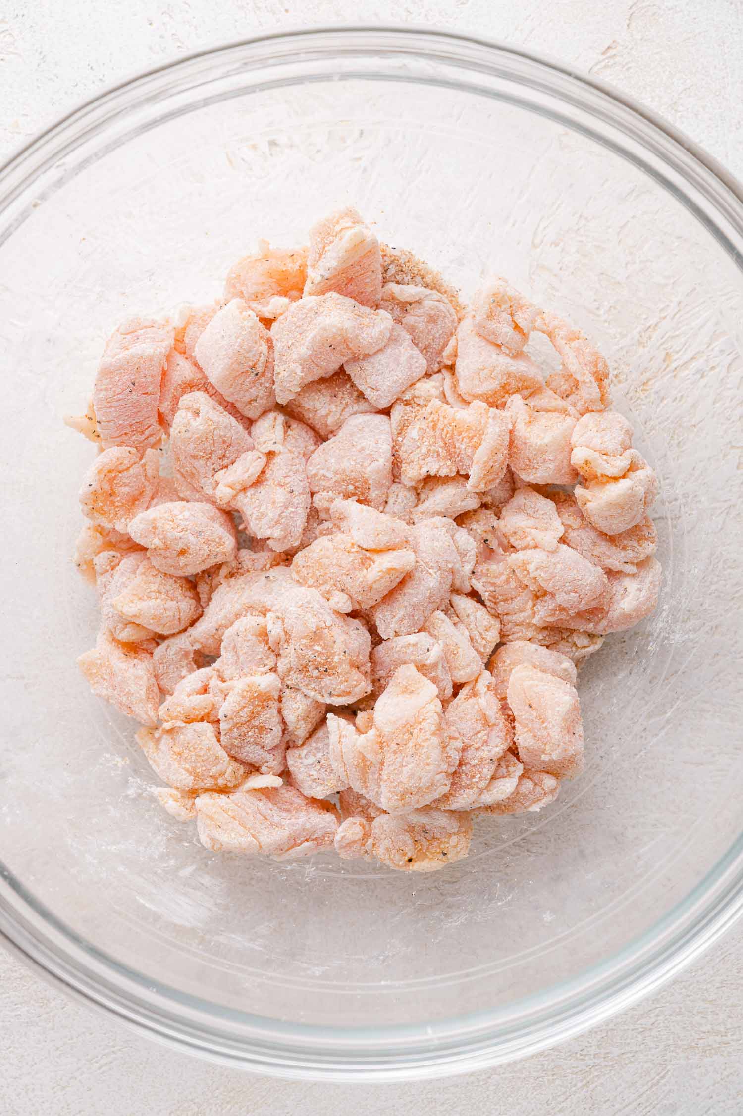 Raw, cubed chicken pieces coated in a light layer of cornstarch and seasonings in a clear glass mixing bowl on a white surface.