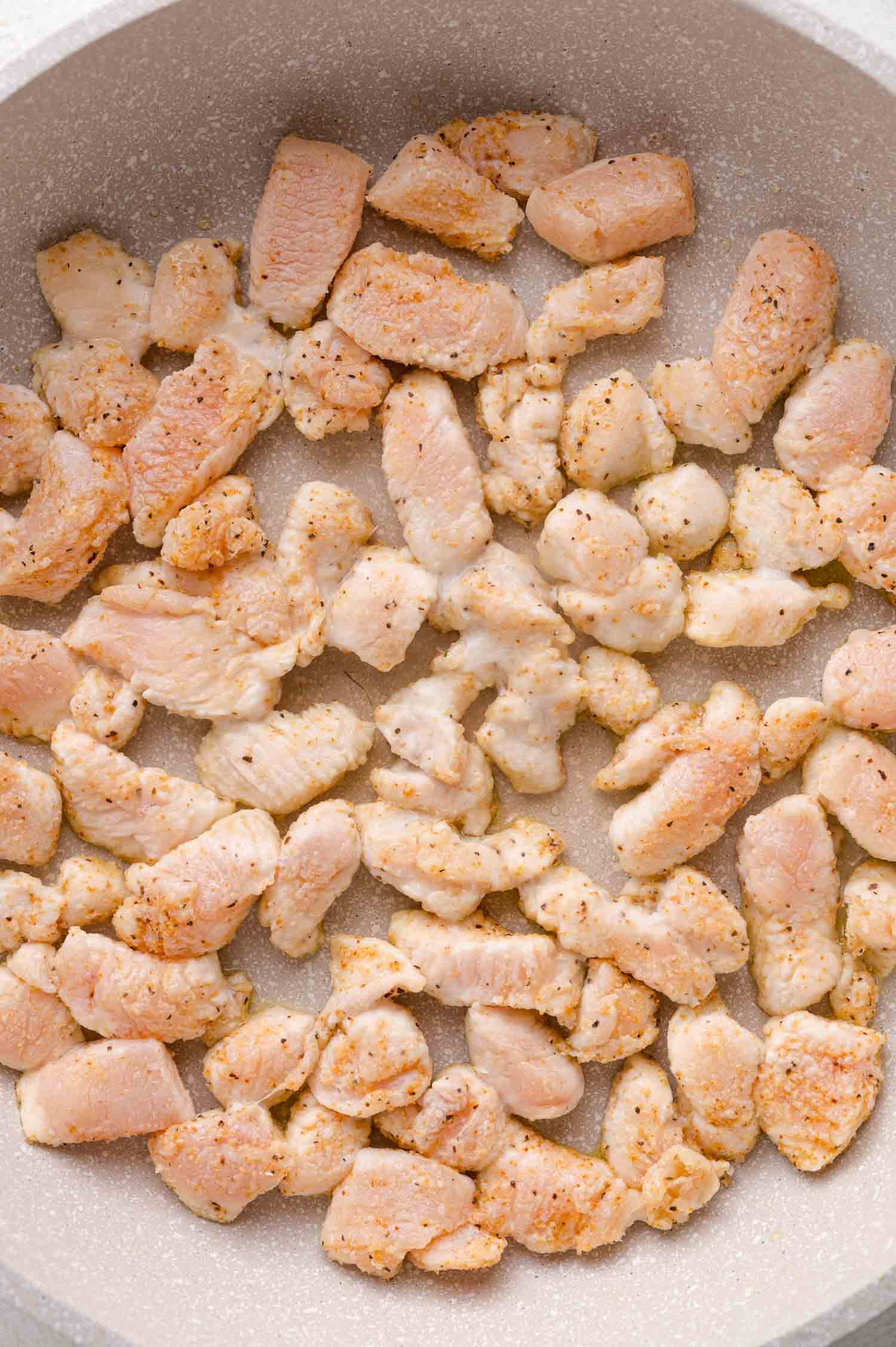 Cubed pieces of seasoned chicken breast cooking in a nonstick pan, lightly browned on the edges.