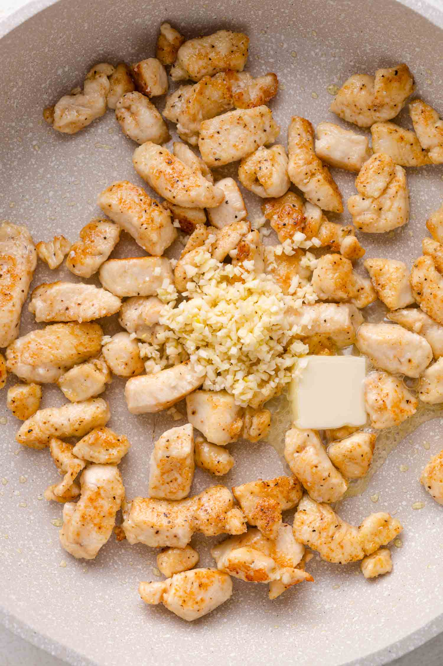 Cubed chicken pieces cooking in a pan with finely chopped garlic and a square of butter in the center.
