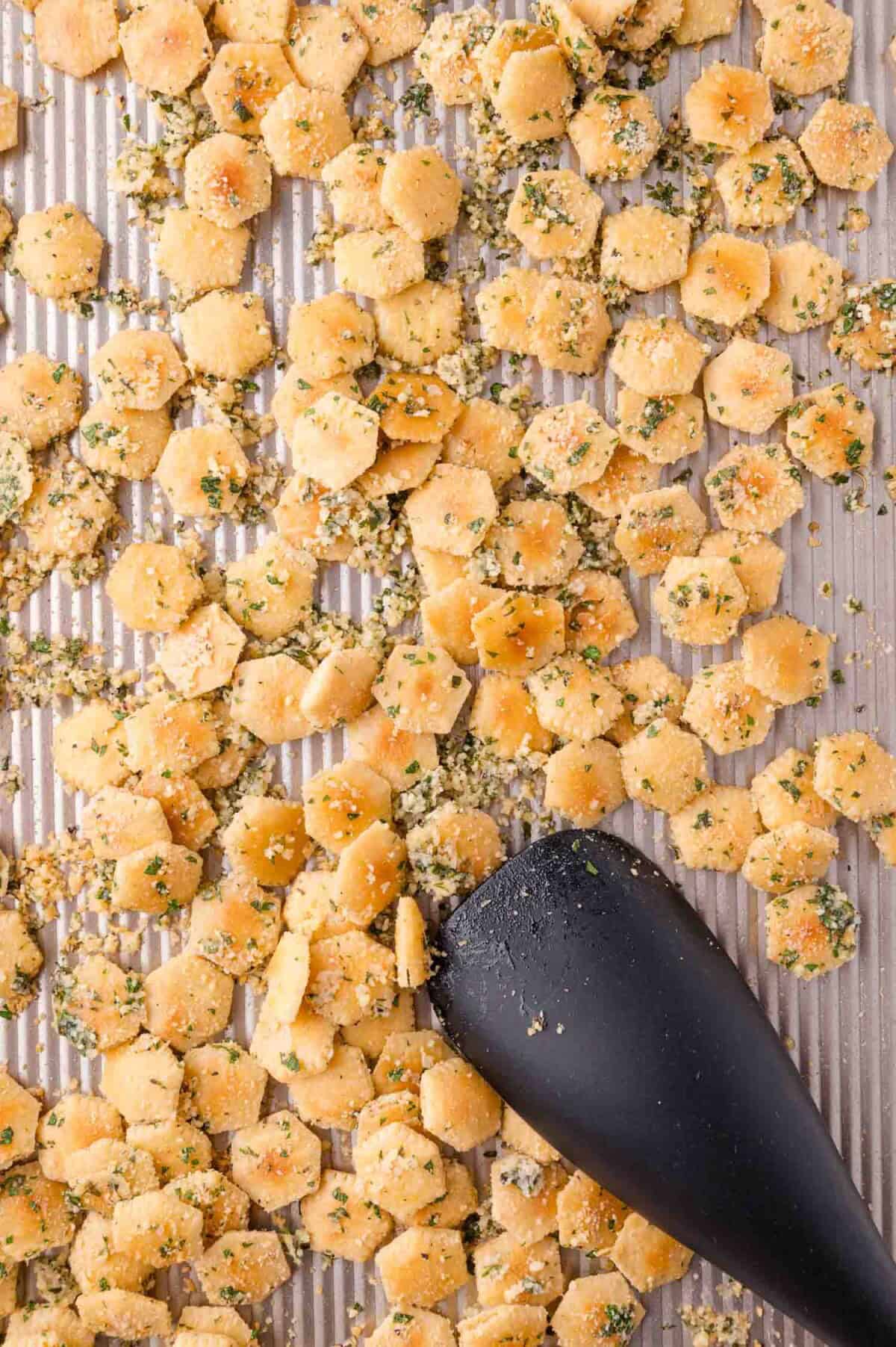 Baked seasoned oyster crackers spread on a ribbed baking sheet, with a black spatula partially visible in the lower right corner.