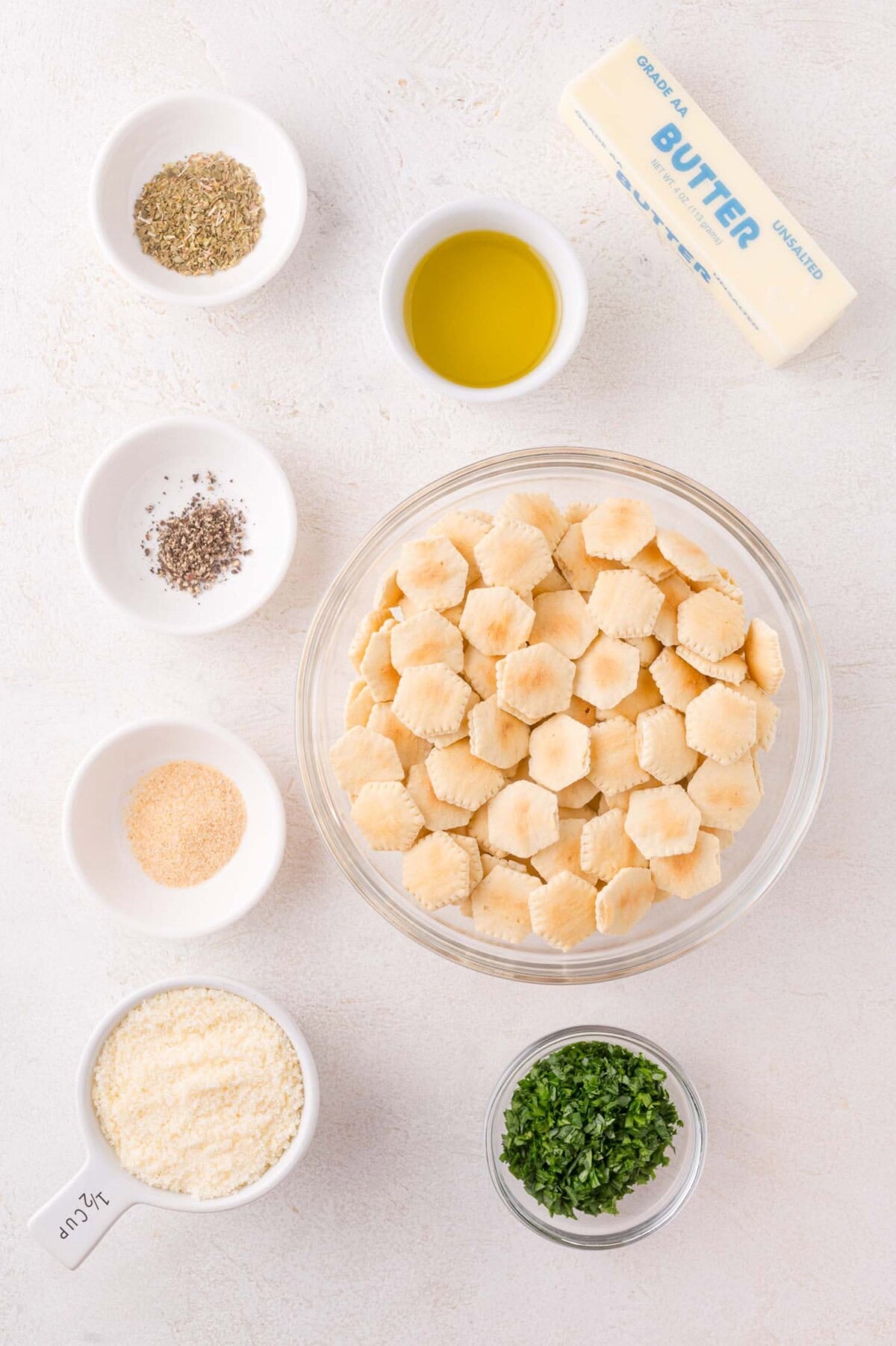 A bowl of oyster crackers is surrounded by small bowls of herbs, spices, grated cheese, olive oil, a stick of butter, and chopped parsley on a white surface.