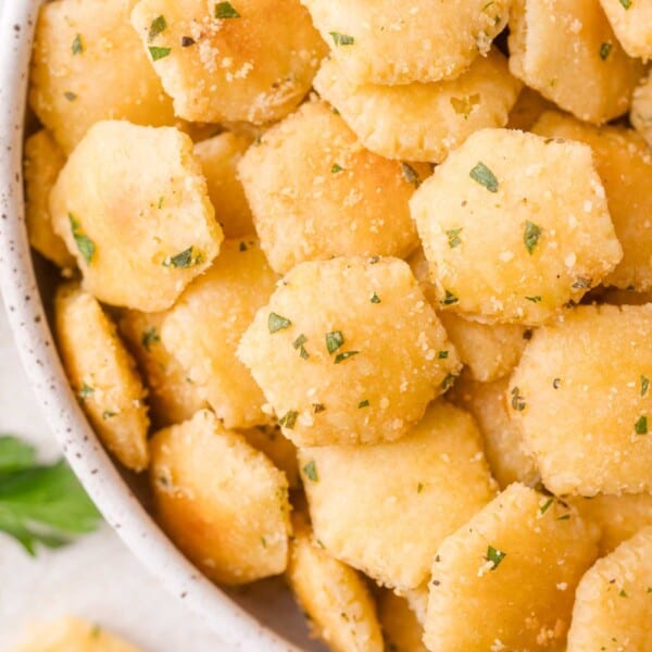 A close-up of a bowl filled with seasoned, hexagonal oyster crackers garnished with herbs.