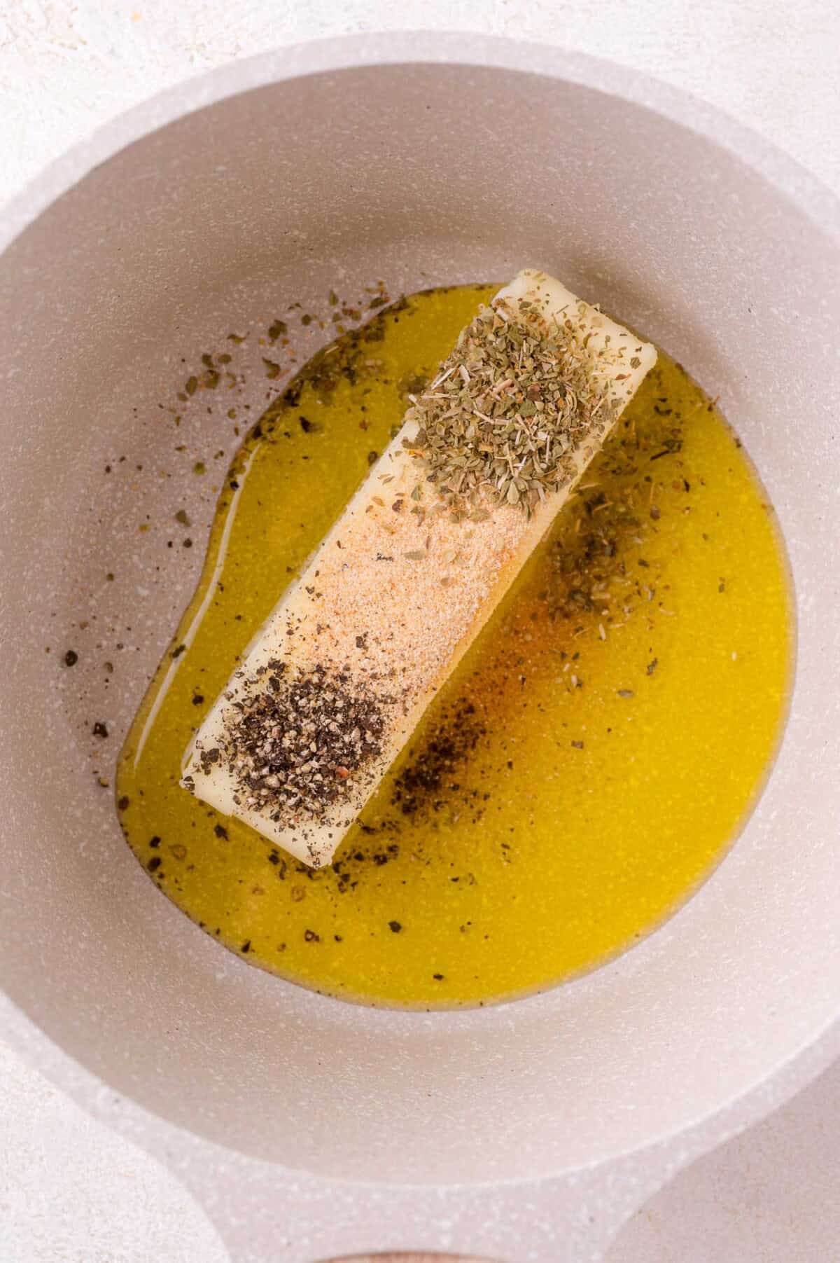 A stick of butter melting in a pan with dried herbs, ground black pepper, and granulated seasoning sprinkled on top.