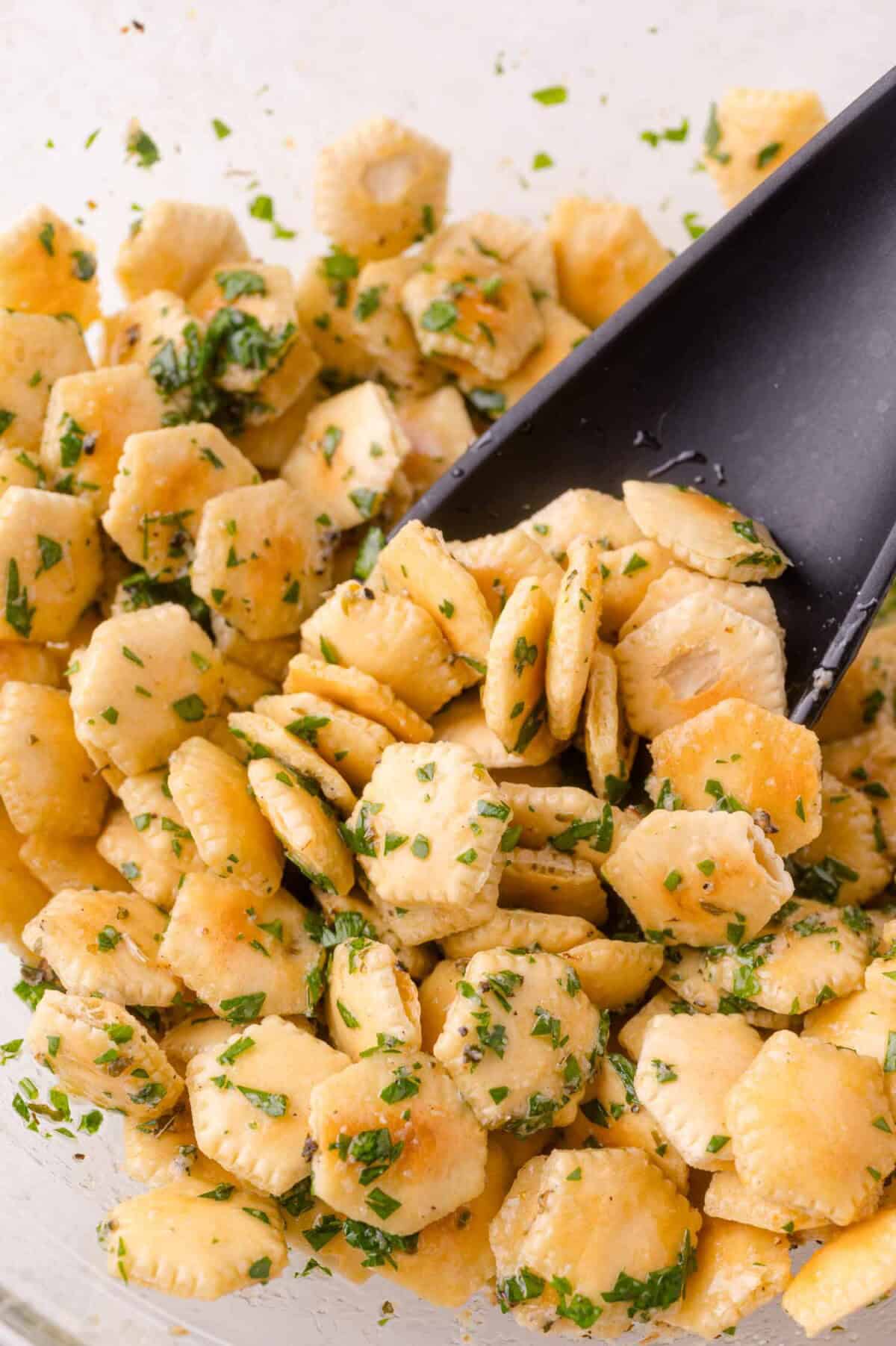 A close-up of seasoned oyster crackers mixed with chopped herbs, being stirred with a black plastic spoon.